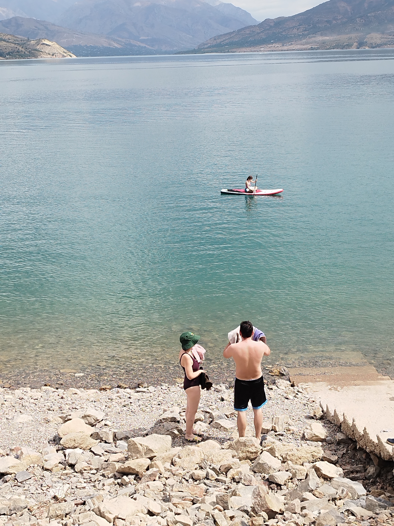 Two people are standing on a rocky shore of a large, calm body of water. The person on the left is wearing a dark swimsuit and a green bucket hat, and has a pink bag slung over their shoulder. The person on the right is a shirtless man wearing black swim trunks. He is drying himself with a white towel. In the distance, a single person is on a paddleboard in the water. The background shows a mountainous landscape under a hazy sky. The location is the Zarafshan River near Yubileiniy, Uzbekistan.