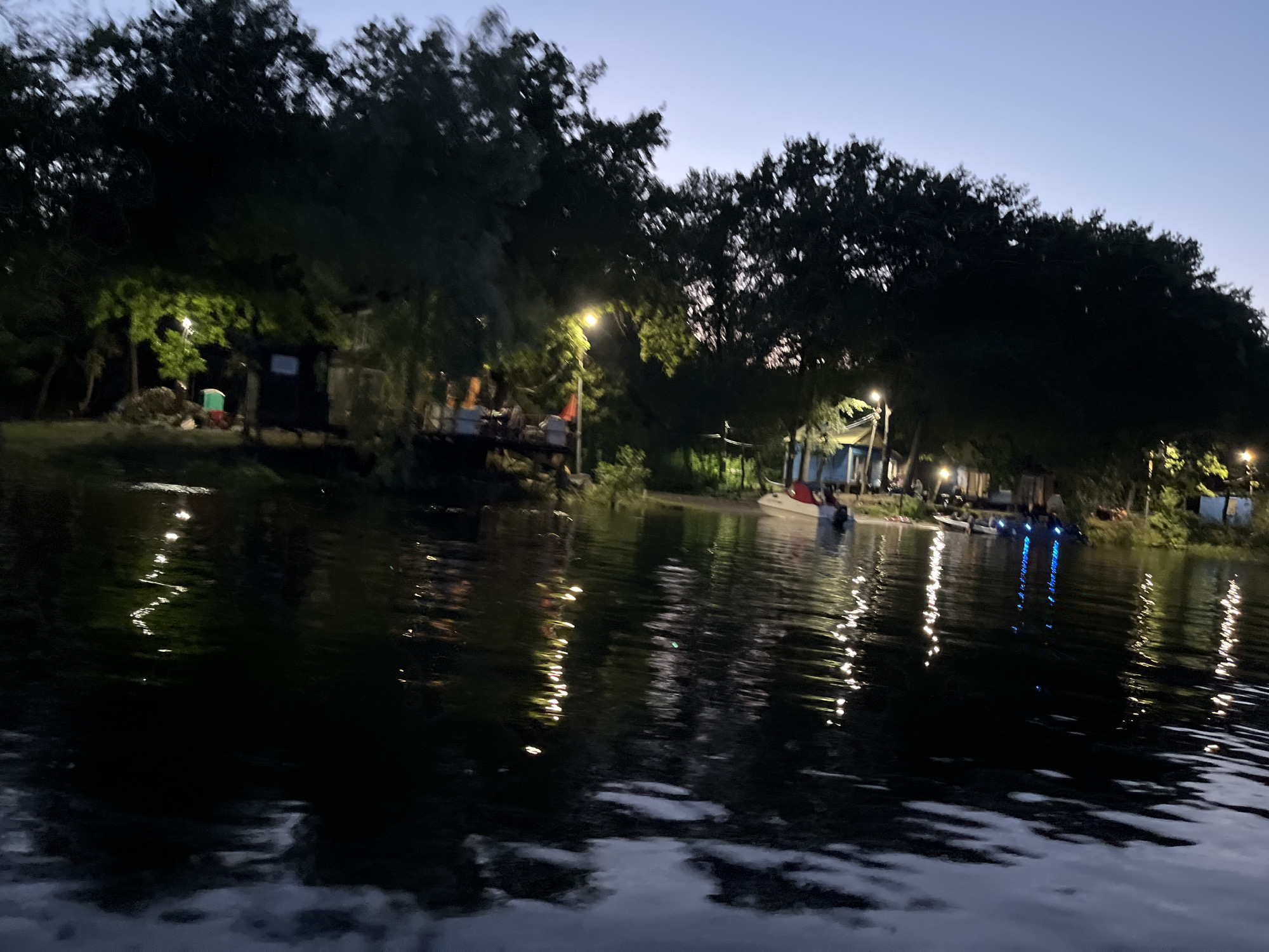 The image depicts a twilight scene at a waterfront location, likely a recreational area or campsite. The sky is a pale blue transitioning to darker shades, indicating it is either dusk or dawn. The foreground is dominated by dark, reflective water, with the lights from the shore creating elongated, shimmering reflections.
Along the shore, numerous trees form a dark silhouette against the sky. There are several structures visible, including what appear to be small buildings or cabins, some with lights on. A portable toilet is visible near one of the structures, illuminated by a bright overhead light. Several boats are moored or docked along the water's edge. Some of these boats have lights on, with blue lights clearly visible reflecting in the water. There are also string lights or other decorative lighting visible along the shore, contributing to the overall ambiance.
The area appears to be a place for recreation or possibly a small village or campsite situated by a body of water. The presence of boats, lights, and structures suggests a populated area, possibly a resort or a private retreat. The overall impression is one of a peaceful, illuminated evening by the water. The specific location in Saratov, Russia, is not discernible from the visual elements within the image itself.
