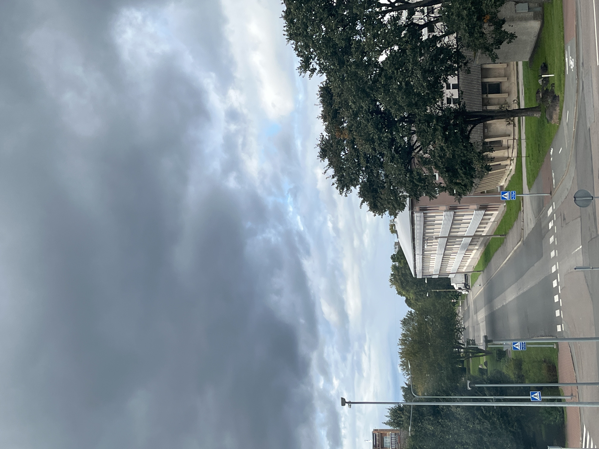 A high-angle, eye-level, full shot captures an overcast day in Kohtla-Järve linn, Estonia. The sky is filled with dark, ominous clouds, with lighter patches of blue peeking through. In the foreground, a street runs diagonally from the bottom left to the upper right, bordered by sidewalks and grassy areas. Several tall, grey lampposts are positioned along the street. A pedestrian crossing is marked on the road. Two blue signs with white symbols indicating pedestrian and bicycle crossings are visible on lampposts.
To the left of the street, a multi-story building with a modern facade and balconies is partially visible. Further down the street, a white truck is parked. On the right side of the street, a long, multi-story building with a light-colored facade and numerous windows stretches into the distance. Large, mature trees with dark green foliage are situated in front of and around the buildings, some obscuring parts of the structures. The overall scene depicts an urban environment under a moody sky.