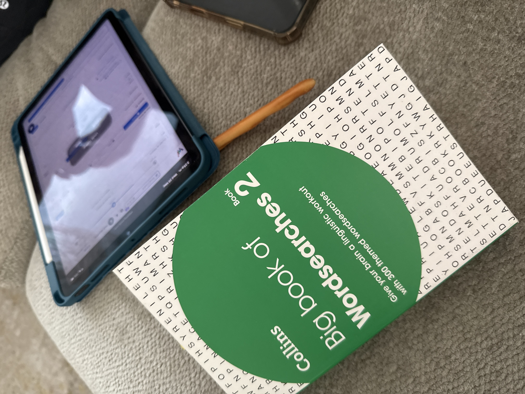 The image shows a still life composition on a gray upholstered surface. Dominating the scene is a "Collins Big book of Wordsearches 2," featuring a green circular label with the book's title in white lettering. The book's corner displays a grid of letters, typical of word search puzzles. An iPad, encased in a teal-colored protective cover, rests partially atop the word search book. An Apple Pencil is attached magnetically to the iPad's side. A smartphone lies behind the book and iPad. The focus is on the book and electronic devices, suggesting leisure activities. There is no information in the image about San Bernardino Tlaxcalancingo, Mexico.