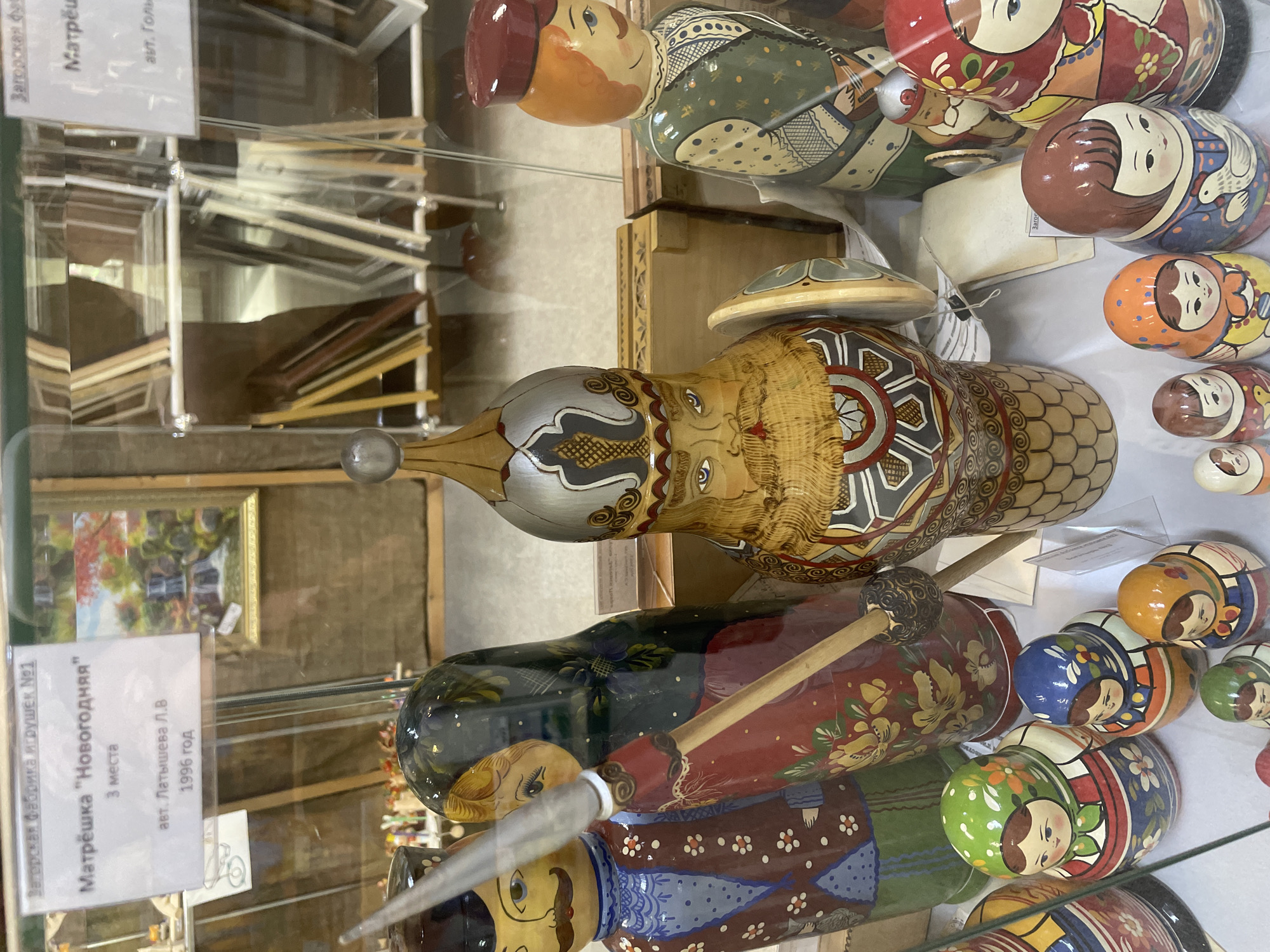 The image shows a display of Russian nesting dolls (matryoshka dolls) in a glass case, likely at a museum or shop in Sergiev Posad, Russia, a city known for its traditional crafts, particularly matryoshka doll production.
In the foreground, several matryoshka dolls are visible. On the left, there is a large matryoshka doll with a blue jacket adorned with red and white floral patterns, a black hat, and a mustache. Next to it is a larger matryoshka doll with a dark green headscarf with floral designs, wearing a red garment, and holding a wooden staff. In the center, a prominent matryoshka doll is depicted with a warrior-like helmet and a stern facial expression, holding a shield. To the right of this central figure is another matryoshka doll with an orange hat and a green vest with dotted patterns.
Smaller matryoshka dolls are arranged in the lower part of the display, featuring colorful traditional patterns on their headscarves and dresses. Some of these smaller dolls have green, blue, and orange headscarves, and are dressed in various floral motifs.
In the background, through the glass, more matryoshka dolls and various wooden items, possibly framed artwork or decorative boxes, can be seen on shelves. A white label with Russian text is attached to the glass, providing information about one of the dolls, including "Матрёшка 'Новогодняя'" (Matryoshka "New Year's"), "3 места" (3 places), and "авт. Латышева Л.В. 1996 год" (auth. Latysheva L.V. 1996). Another label, partially visible, mentions "Загорская фабрика игрушек №1" (Zagorsk Toy Factory No. 1), referring to the historical name for Sergiev Posad and its famous toy factory. The lighting suggests an indoor environment, likely a display case in a retail or museum setting.