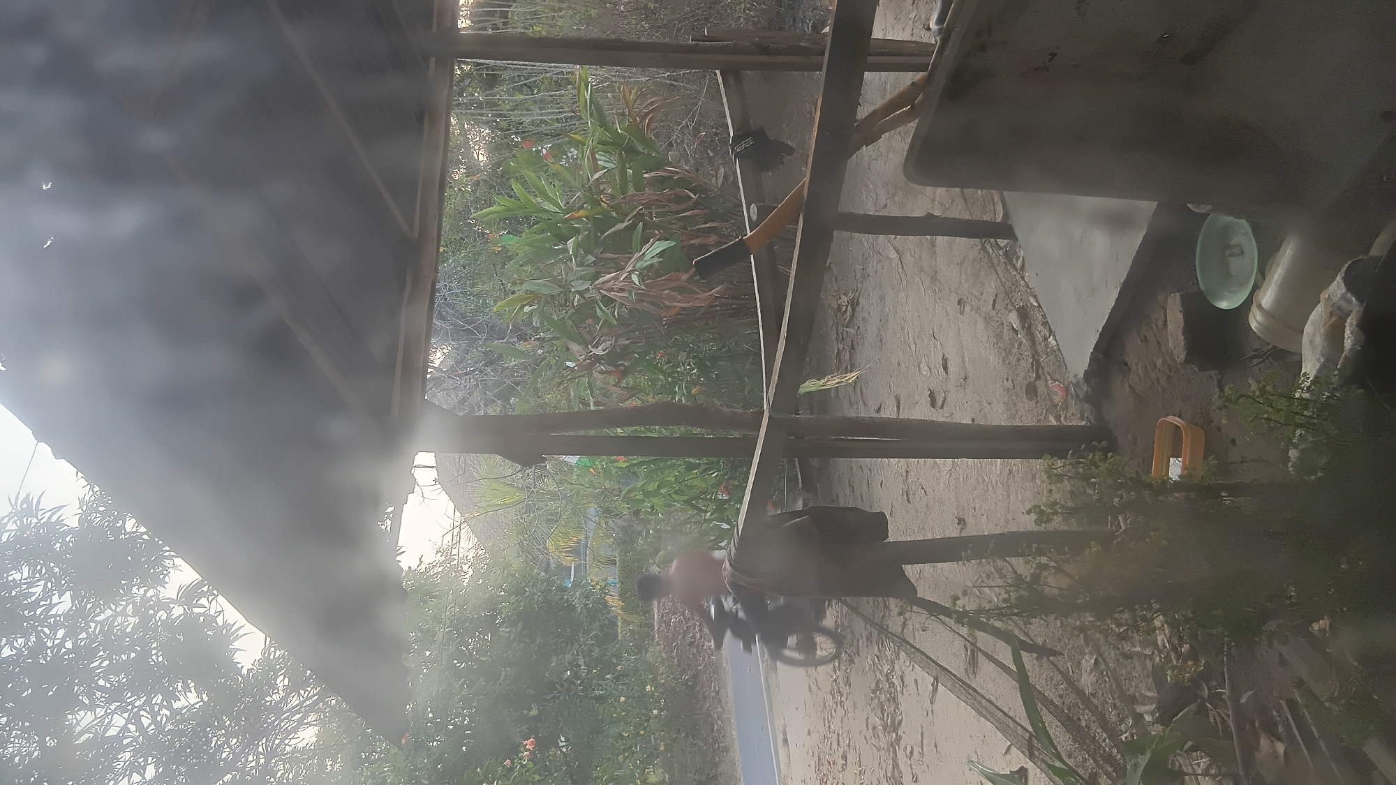 A person is riding a motorcycle on a dirt road, partially obscured by trees and foliage. The scene appears to be an outdoor rural or semi-rural setting, possibly in Prabumulih, Indonesia, given the context. A weathered, open-sided structure with a corrugated metal roof is in the foreground. A dark colored garment hangs from a wooden beam within the structure. The ground is sandy and covered with leaves. The overall atmosphere is hazy, possibly due to humidity or dust.