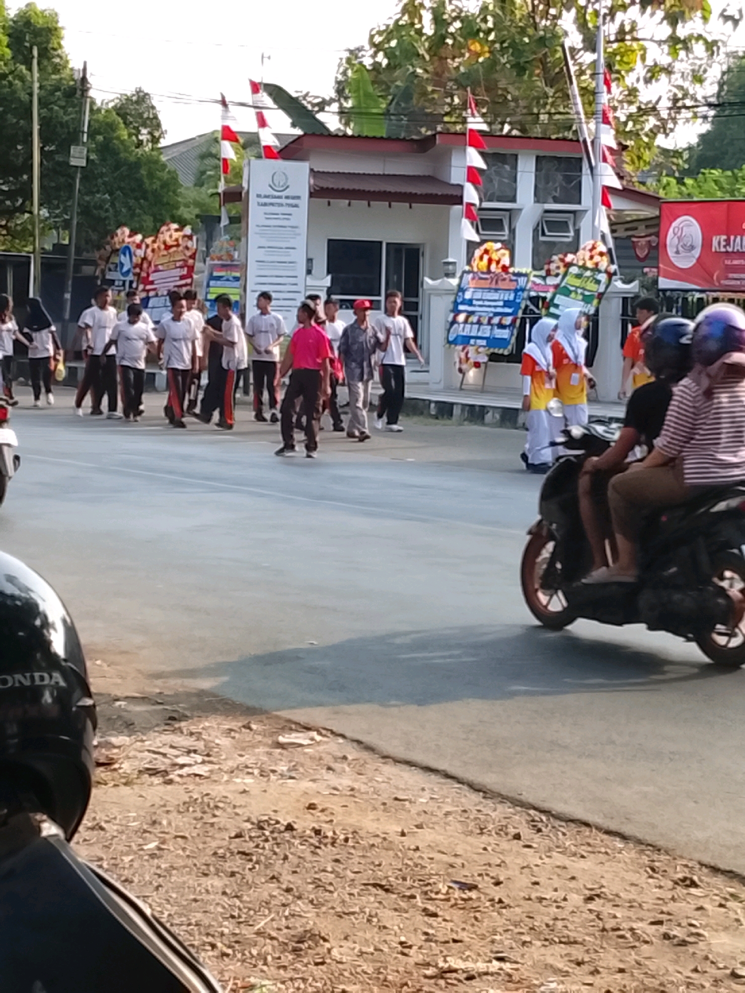 The image depicts a street scene in Slawi, Indonesia, likely during a daytime event. A group of people, primarily young males wearing white shirts and dark pants, are walking on the road. Some are facing the camera, while others are walking away. A person in a bright pink shirt is walking towards the right side of the frame, while another in a red shirt is partially visible behind them. Two individuals, a man and a woman, are riding on a black scooter in the foreground, moving from right to left. The woman is wearing a striped shirt and a hat, and the man is wearing a black shirt. In the background, a building with red and white flags on poles is visible, along with banners and decorations. The overall atmosphere appears to be lively, possibly a parade or a community gathering. The road is paved, and the area alongside it is dirt with scattered debris. The lighting suggests it is a sunny day.