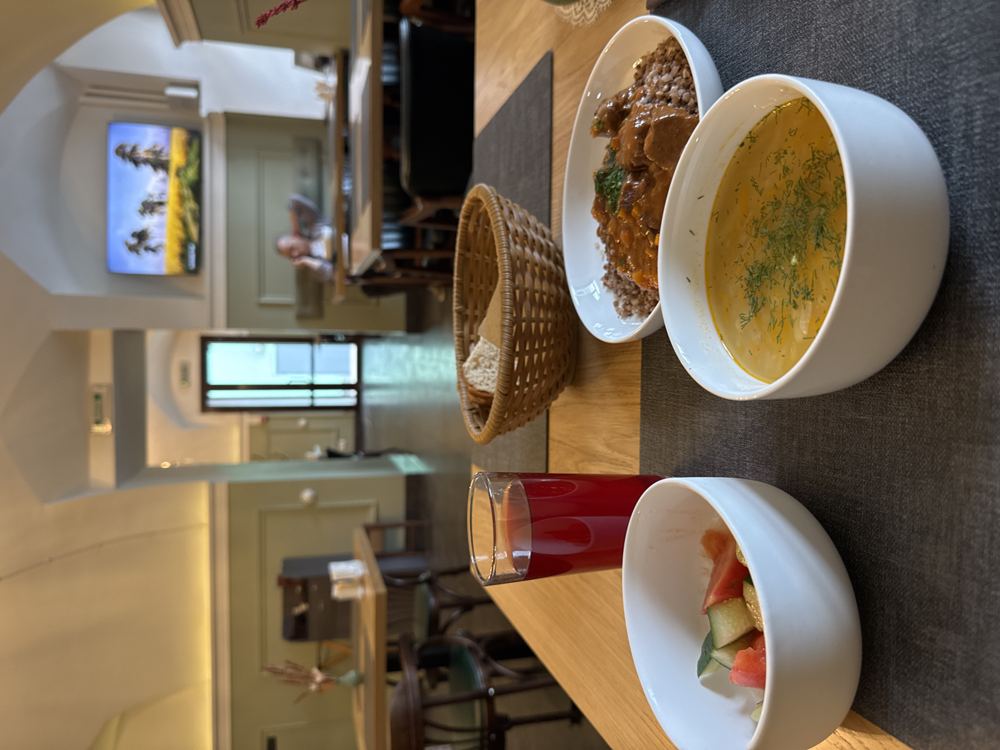 A close-up shot captures a meal laid out on a wooden table in what appears to be a restaurant. In the foreground, three white bowls are arranged. The leftmost bowl contains a chopped salad with visible pieces of tomato and cucumber. Adjacent to it, a bowl of soup is filled with a yellowish broth and garnished with dill. The third bowl, positioned further back, holds a dish of buckwheat topped with a rich, brown stew. A glass of red liquid, likely juice or compote, stands beside the salad bowl. A woven basket filled with bread sits to the right of the drink. The background reveals a dining hall with tables and chairs, and a television mounted on the wall displaying a landscape scene. One person is visible in the background, seated at a table and appearing to be on their phone. The ambiance suggests a casual dining establishment, possibly a cafeteria or a more budget-friendly eatery. The overall scene is brightly lit, indicating daytime. The presence of Cyrillic lettering on signs in the background suggests the location is within Russia, likely Moscow due to the commonality of such establishments in the city.