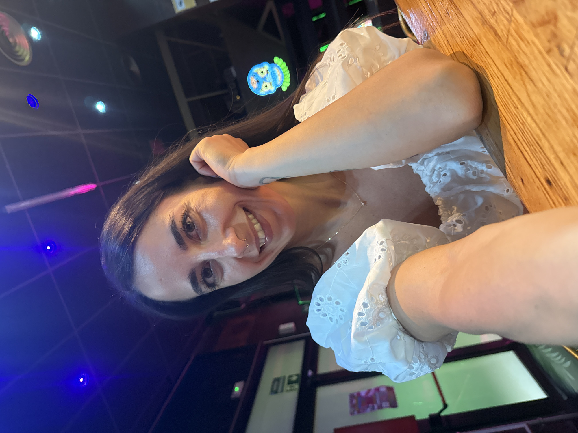 A medium shot captures a woman with long dark hair, smiling and resting her chin on her hand, as she takes a selfie. She is wearing a white eyelet top with puff sleeves and a delicate necklace with a small diamond pendant. Her left arm is extended towards the camera, and her right elbow rests on a wooden surface that appears to be a bar. Behind her, the environment is dimly lit with colorful neon lights from a sign that depicts a blue-faced, green-haired creature. The background also shows a door with a green exit sign and a hint of what looks like a bar interior. The lighting suggests the photo was taken indoors at night.