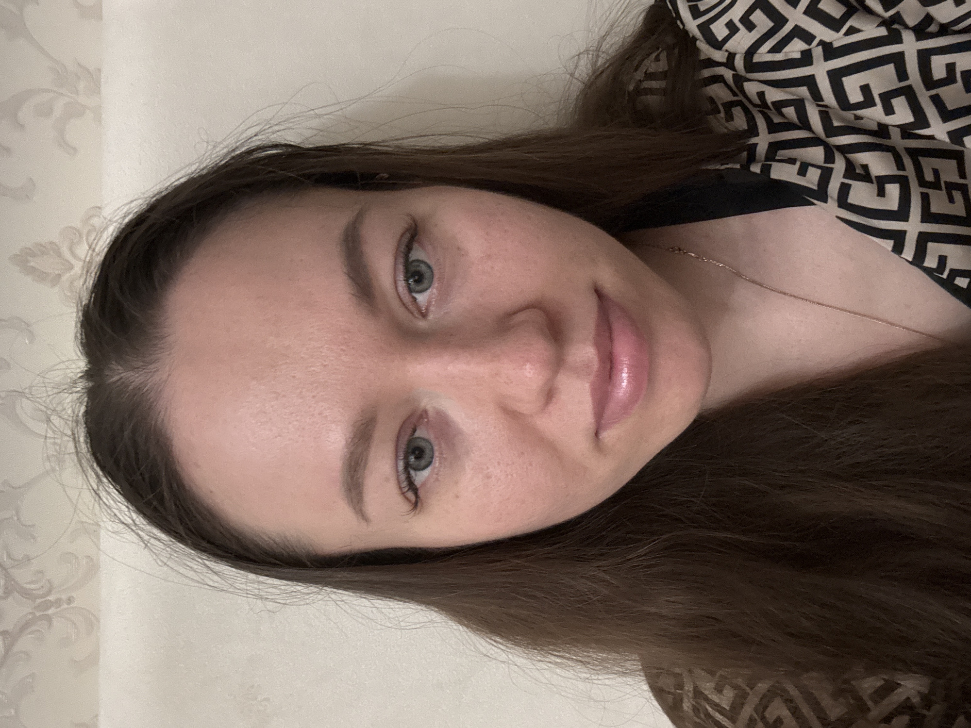 A young woman is pictured from a close, slightly upward angle. She has fair skin, light eyes, and brown hair. She is wearing a top with a geometric print and a thin gold necklace. The background shows a light-colored wall with a subtle floral pattern. The location is not determined.