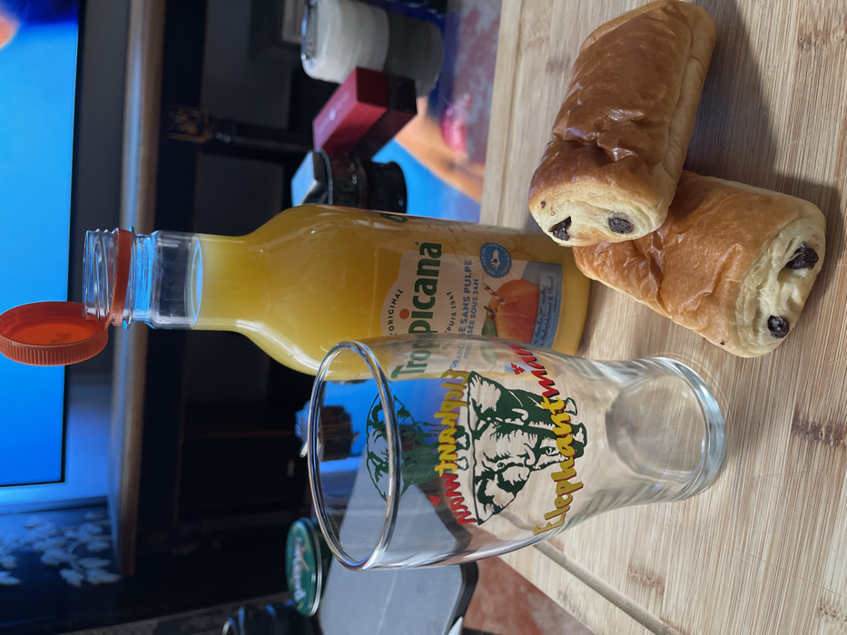 The image depicts an indoor scene focused on a prepared breakfast or snack on a light-colored wooden cutting board. Two golden-brown *pains au chocolat* (chocolate croissants), one slightly overlapping the other, rest on the board. To their left is an empty clear glass tumbler adorned with a green graphic of two elephant heads and the yellow/red text "Elephantman". Behind these items stands a clear plastic bottle of Tropicana Original orange juice, featuring an open orange flip-top cap. The label explicitly states "Jus de fruits sans pulpe" (fruit juice without pulp), indicating a French-speaking region.
The background, largely out of focus, reveals an entertainment unit or shelf beneath a large screen, likely a television, which emits a prominent blue light. Various objects are visible on the shelf, including what appears to be a dark, possibly ornate metal object (resembling a hookah component or a lighter), a rectangular red object (possibly a box), a white translucent container, and a round green-lidded tin. The environment suggests a domestic setting within France. Specific visual identifiers to precisely locate the scene within the city of Plaisir are not present in the image.