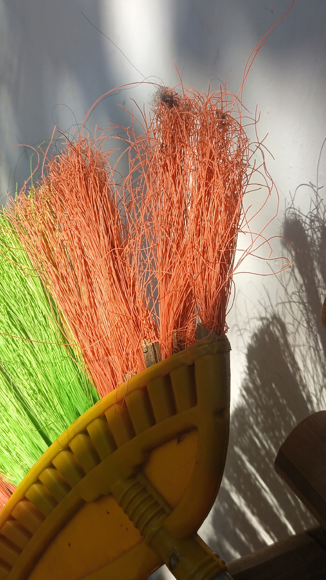 A close-up shot shows the head of a broom with green bristles on the left and orange bristles on the right. The broom head is made of yellow plastic with a ribbed texture. The bristles are uneven and some have fallen out, with a few dark fibers and a small piece of debris visible among the orange bristles. The broom is positioned against a white wall that is partially illuminated by sunlight, casting shadows of the bristles onto the wall. The scene appears to be outdoors in Bulungcangkring, Indonesia, given the natural lighting and the texture of the wall which is common in Indonesian houses. There are no people or animals visible in the image.