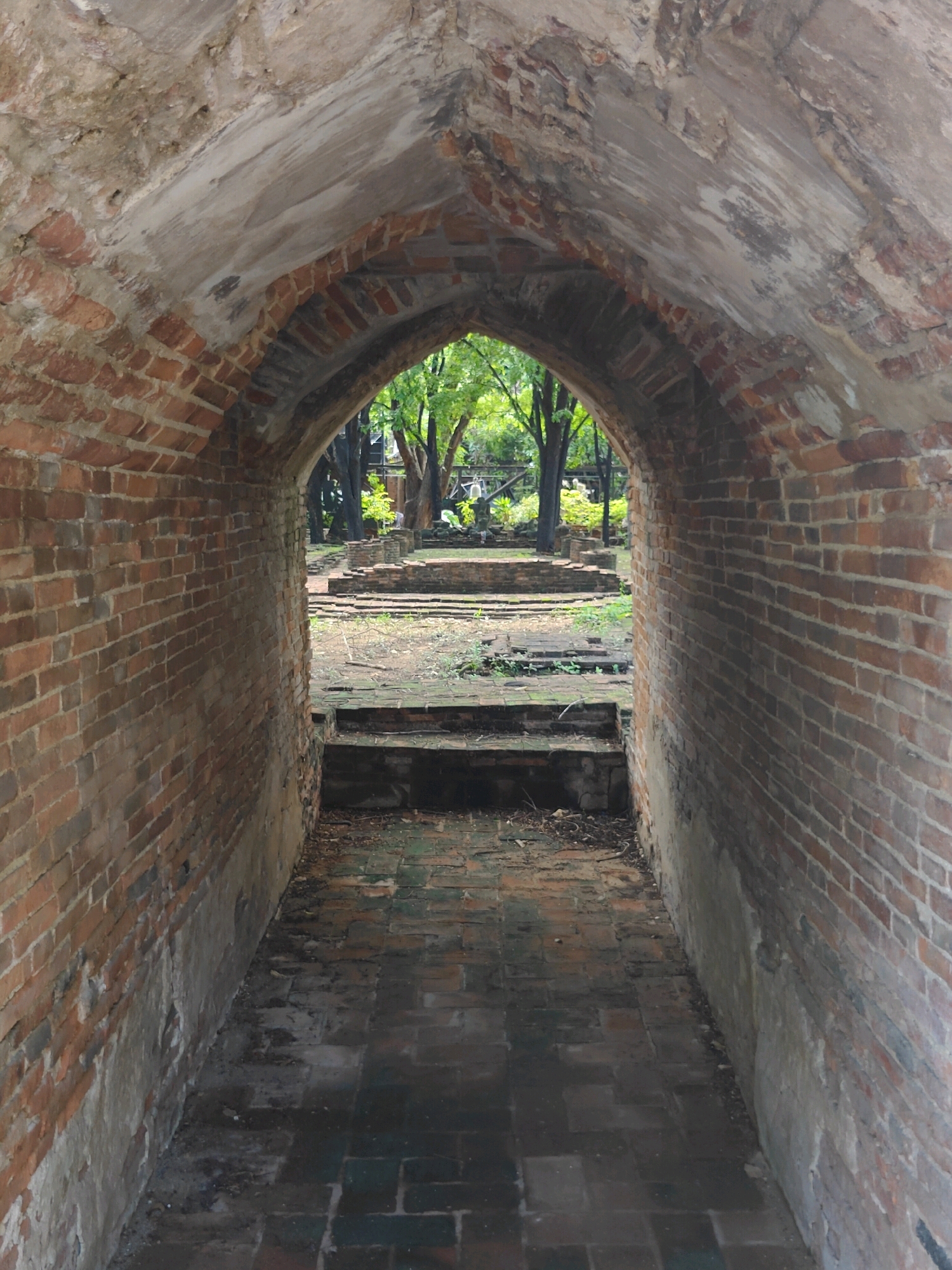 The image shows an arched brick tunnel leading to an outdoor area with trees and what appears to be ruins. The tunnel walls are constructed of red bricks, some of which are weathered and chipped. The ceiling of the tunnel is also bricked and arched, with some areas showing exposed plaster or cement. The floor of the tunnel is paved with dark bricks. Outside the tunnel, there are stone steps and remnants of old structures visible amidst greenery. The overall impression is of an ancient, possibly historical site, likely located in a humid, tropical environment, consistent with the city of Phra Nakhon Si Ayutthaya in Thailand, known for its historical ruins. There are no people or animals visible in the image.