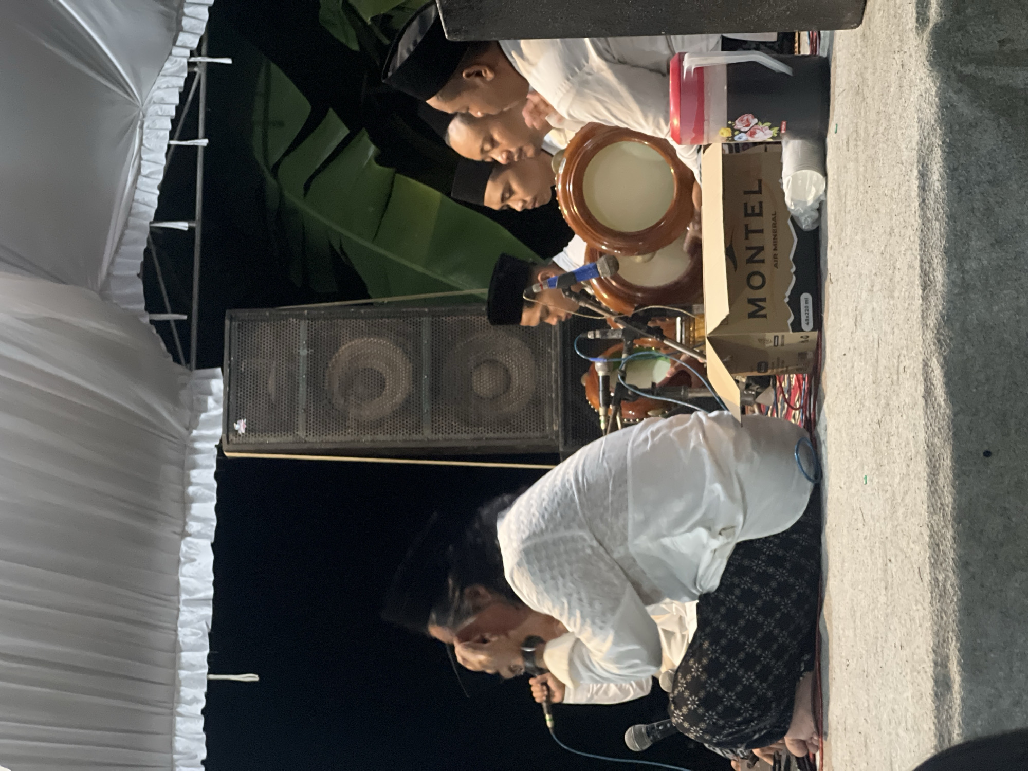 In Wedarijaksa, Indonesia, a group of men dressed in white attire and black skullcaps are seated on a stage under a white tent. The man in the foreground, with long dark hair and a black skullcap, is holding a microphone and appears to be singing or speaking. He is wearing a white, intricately patterned shirt and black patterned pants. To his right, four other men are also seated, three of whom are holding tambourines. Behind them, a large speaker is visible, along with what appears to be banana leaves from the environment. In front of the group, a cardboard box with "MONTEL AIR MINERAL" printed on it and a large jug of dark liquid with a pink lid are placed on the ground. The scene suggests a performance or a gathering with musical accompaniment.