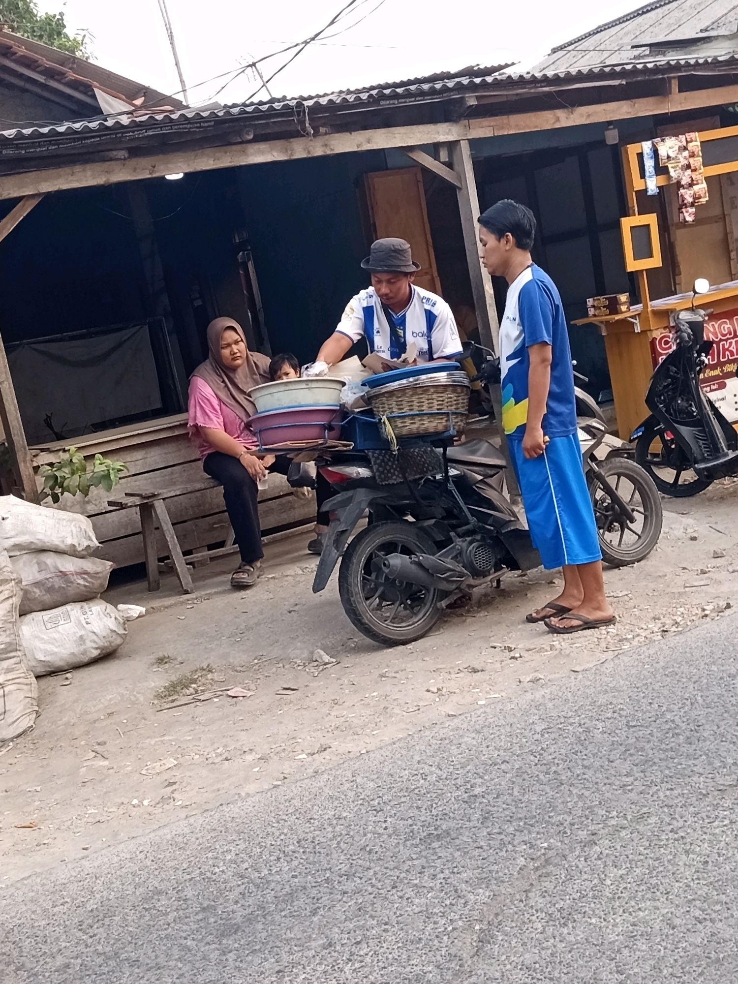 In Karawang, Indonesia, a street scene unfolds in front of a modest, open-air shop with a corrugated metal roof. A woman wearing a hijab and a pink shirt is seated on a wooden bench, holding a young child on her lap. Two young men stand near a parked motorcycle. One man, wearing a blue and white t-shirt and a hat, appears to be serving or interacting with a customer. The other man, in a blue t-shirt and blue shorts, stands passively. The motorcycle is laden with baskets and containers, suggesting it is used for transporting goods. Several large sacks are piled on the ground to the left of the shop. The environment appears to be a roadside, with a paved road in the foreground and a dirt path leading to the shop. The overall impression is one of everyday commerce and street life.