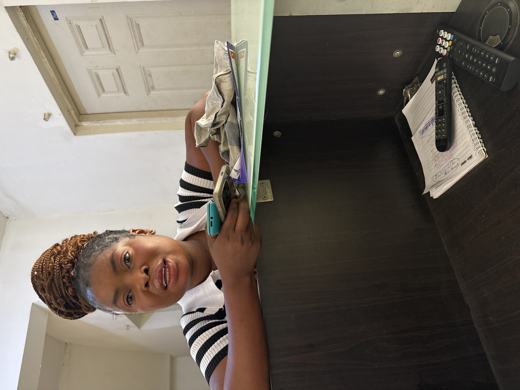 The image shows a Black woman indoors, likely in Abuja, Nigeria, positioned as if looking over a counter or desk. She has brown braided hair styled in an updo and is wearing a white top with black horizontal stripes. She holds a light blue smartphone in her right hand and looks directly at the camera with a neutral to slight smile.
Behind her, to the left of the frame (when upright), is a white paneled door against a light-colored wall, with some small metal fixtures visible. To her right, a dark wooden desk or counter surface holds various items. These include two black remote controls, one featuring colorful buttons, and a spiral-bound notebook containing handwritten notes. A white and black checkered cloth is also visible on the desk. A translucent light green divider separates the woman from the main desk area.
The visible text from the notebook includes numbers and codes such as "95", "105", "51", "0001:", "0000", "000", "231 Total", "ECO-ROUK", "SWORD-SPRIT", and "253/16/2/2". The setting appears to be a casual indoor environment, possibly a home office or reception area.