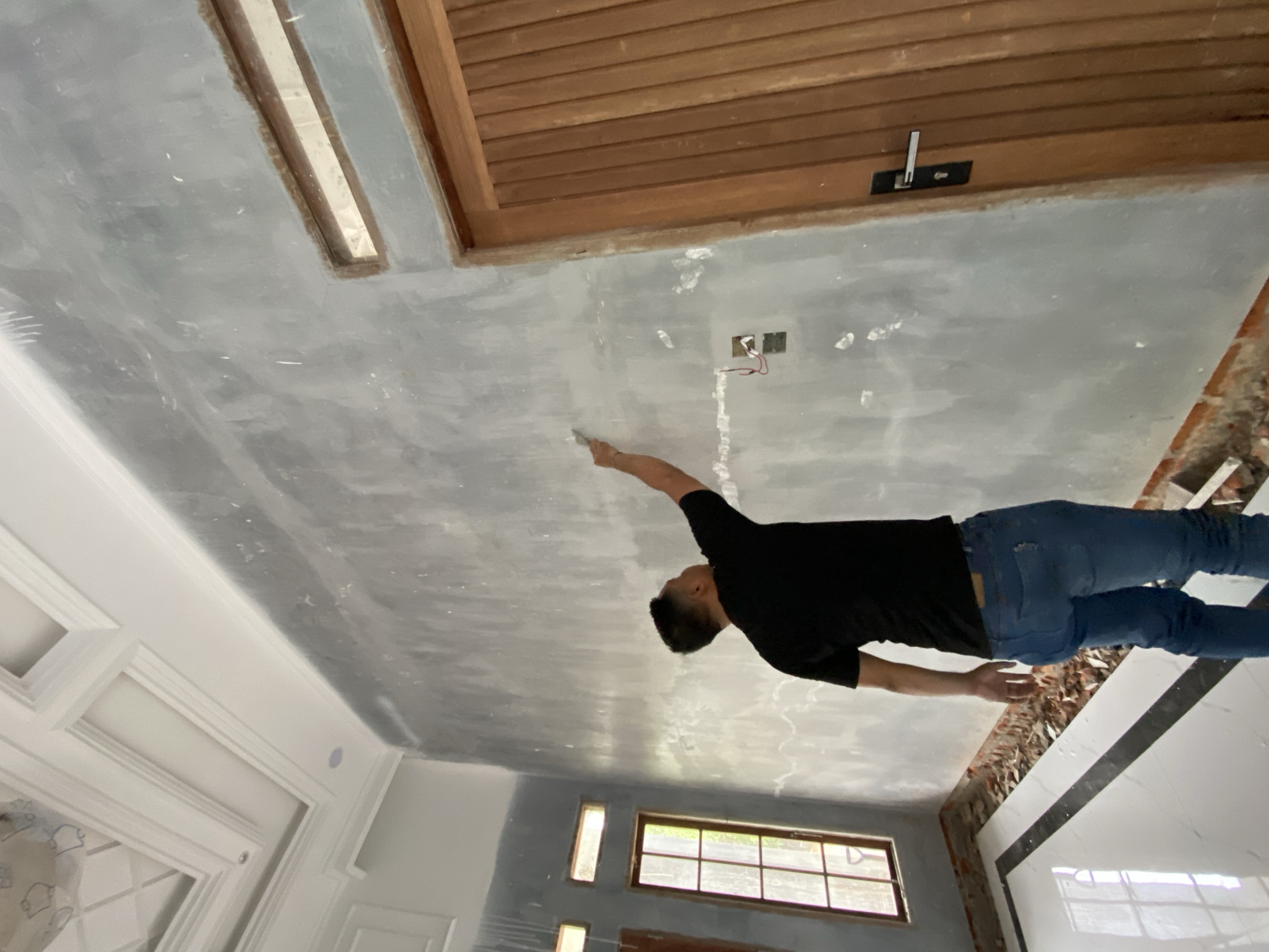 The image shows a man in a black t-shirt and blue jeans applying plaster to a ceiling. He is holding a trowel in his right hand and his arm is extended upwards as he works. The ceiling appears to be a work in progress, with patches of plaster applied over a grey base. The room has unfinished elements, including exposed brickwork and electrical outlets on the ceiling. There are also wooden beams and a wooden slatted ceiling panel visible in the upper part of the image. The room is lit by natural light, suggesting it is daytime. The style of the ceiling and decorative molding on the walls indicates an interior residential or commercial space undergoing renovation or construction. The man's posture and focused action suggest he is engaged in manual labor related to finishing the ceiling. No text is visible in the image.
