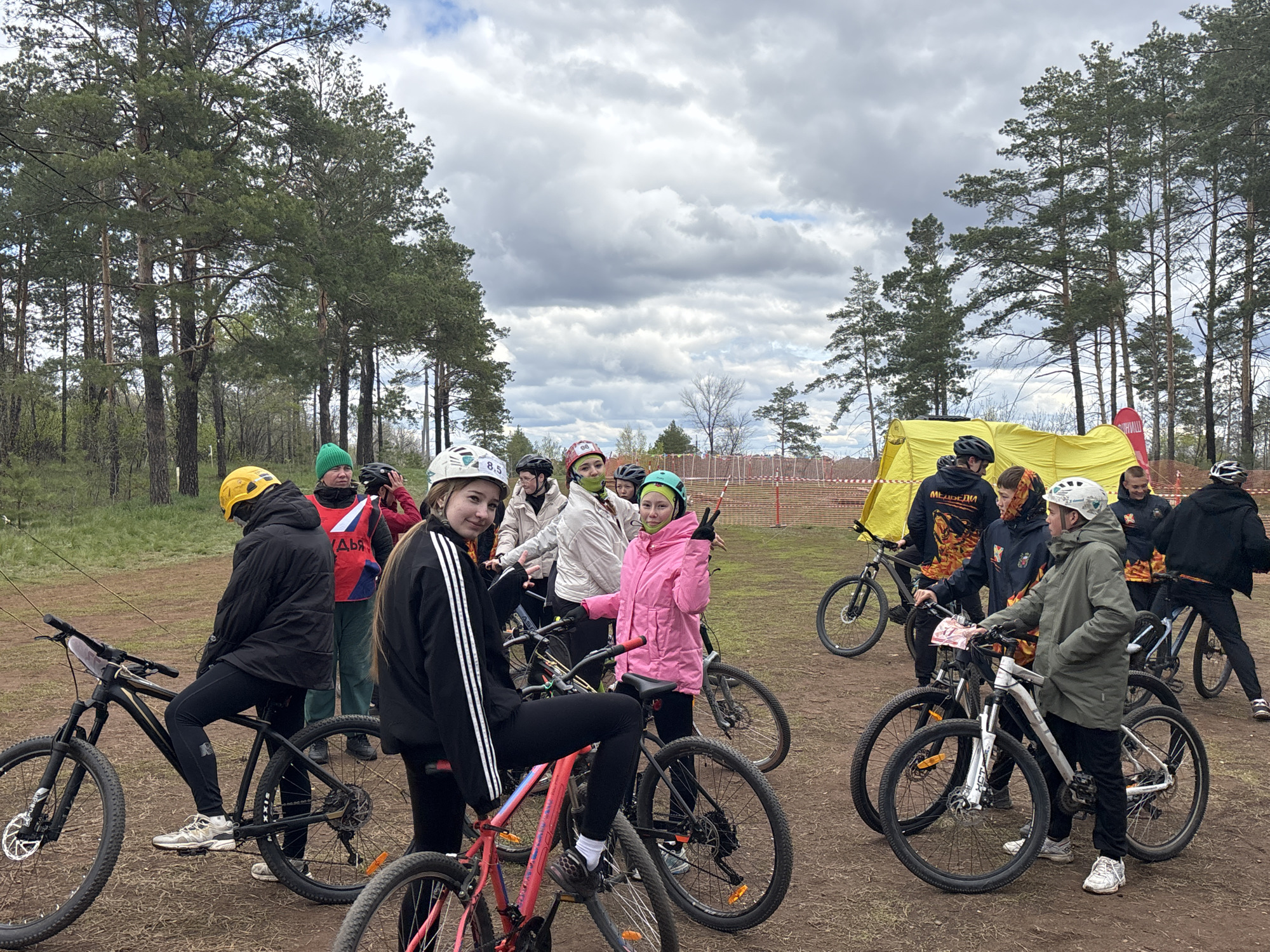 A group of young people and adults are gathered with their bicycles in what appears to be a park or wooded area, likely in the city of Orenburg, Russia. They are dressed in casual attire and athletic gear. Some are wearing helmets. There is a yellow tent with some red flags in the background. Trees are scattered throughout the scene. The ground is dirt and sparse vegetation. The sky is overcast.