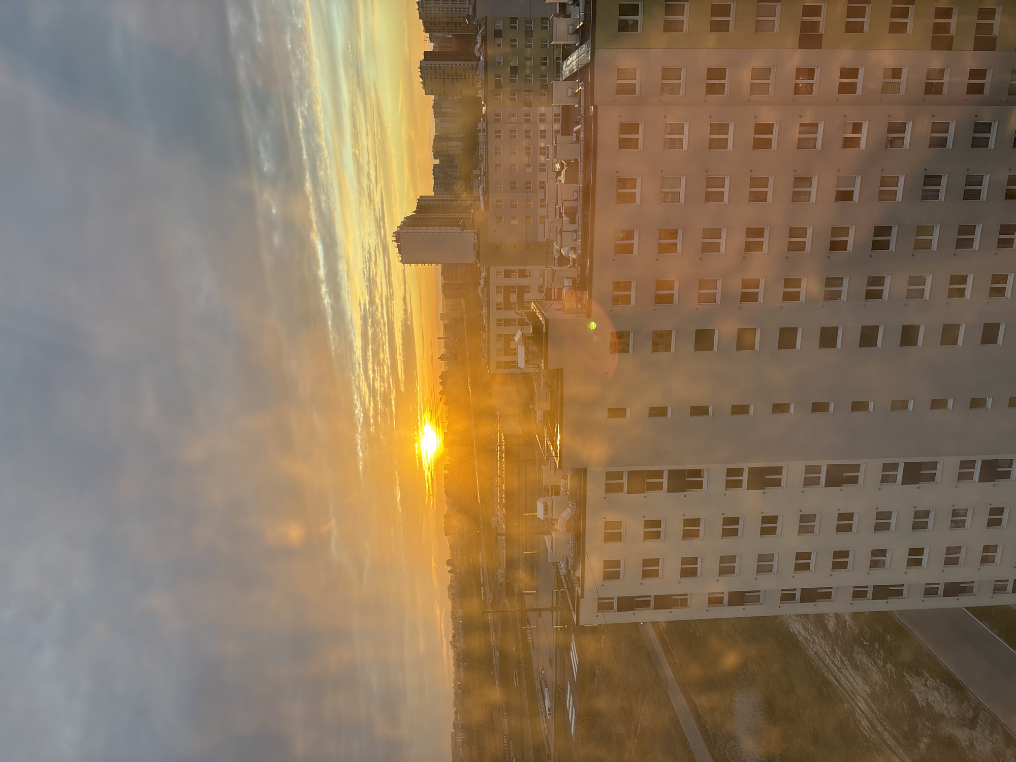 The image depicts a sunrise over Vsevolozhsk, Russia, seen from a high vantage point, possibly an upper floor of a high-rise apartment building. The sun is low on the horizon, casting a strong golden light that illuminates the sky, the buildings, and the surrounding landscape. The sky displays streaks of light breaking through the clouds. There are numerous buildings visible, including a large, light-colored building with many windows, partially dominating the right side of the view. The ground below features a network of roads or paths and some green spaces. The perspective is slightly tilted to the right. There are no visible people.