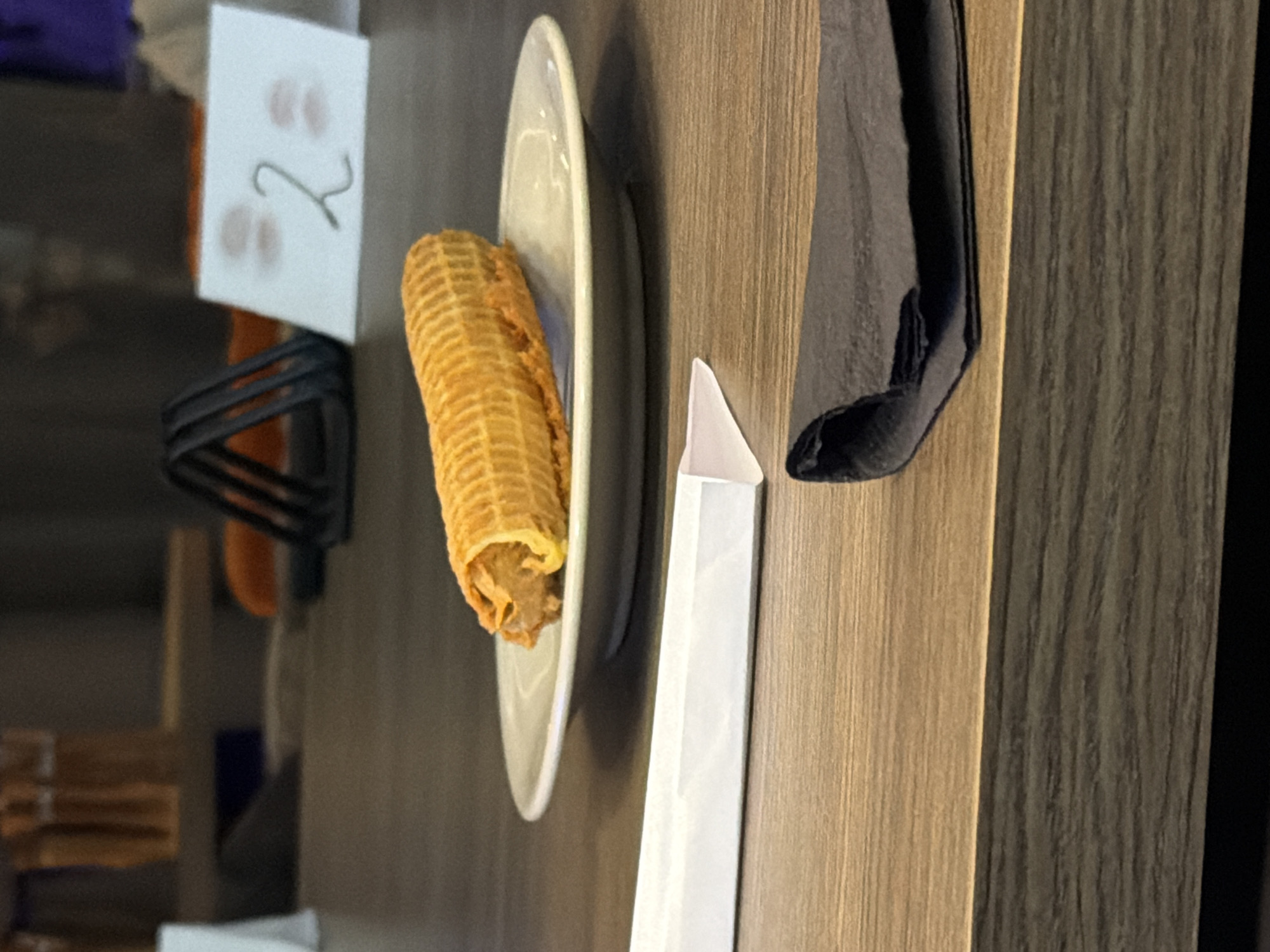 The image shows a table setting with a waffle roll on a white plate, a white napkin, and a black napkin. There is a small white card with the number "2" written on it, standing upright in a holder. The table has a wooden surface with light and dark brown tones. The location is Unknown, Unknown.