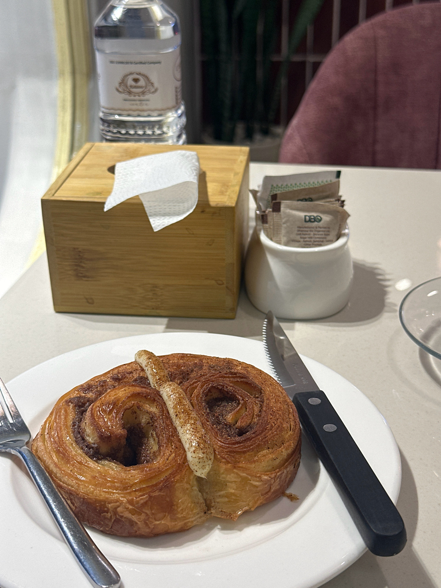 A close-up shot shows a cinnamon roll on a white plate, with a fork to the left and a knife to the right. In the background, a bamboo tissue box with a white tissue sticking out sits on a table, next to a small white ceramic holder filled with sugar packets. A bottle of water is also visible in the background, along with the arm of a pink upholstered chair. The setting appears to be a cafe or restaurant in Kathmandu Metropolitan City, Nepal, likely indoors. The lighting is dim, creating soft shadows.