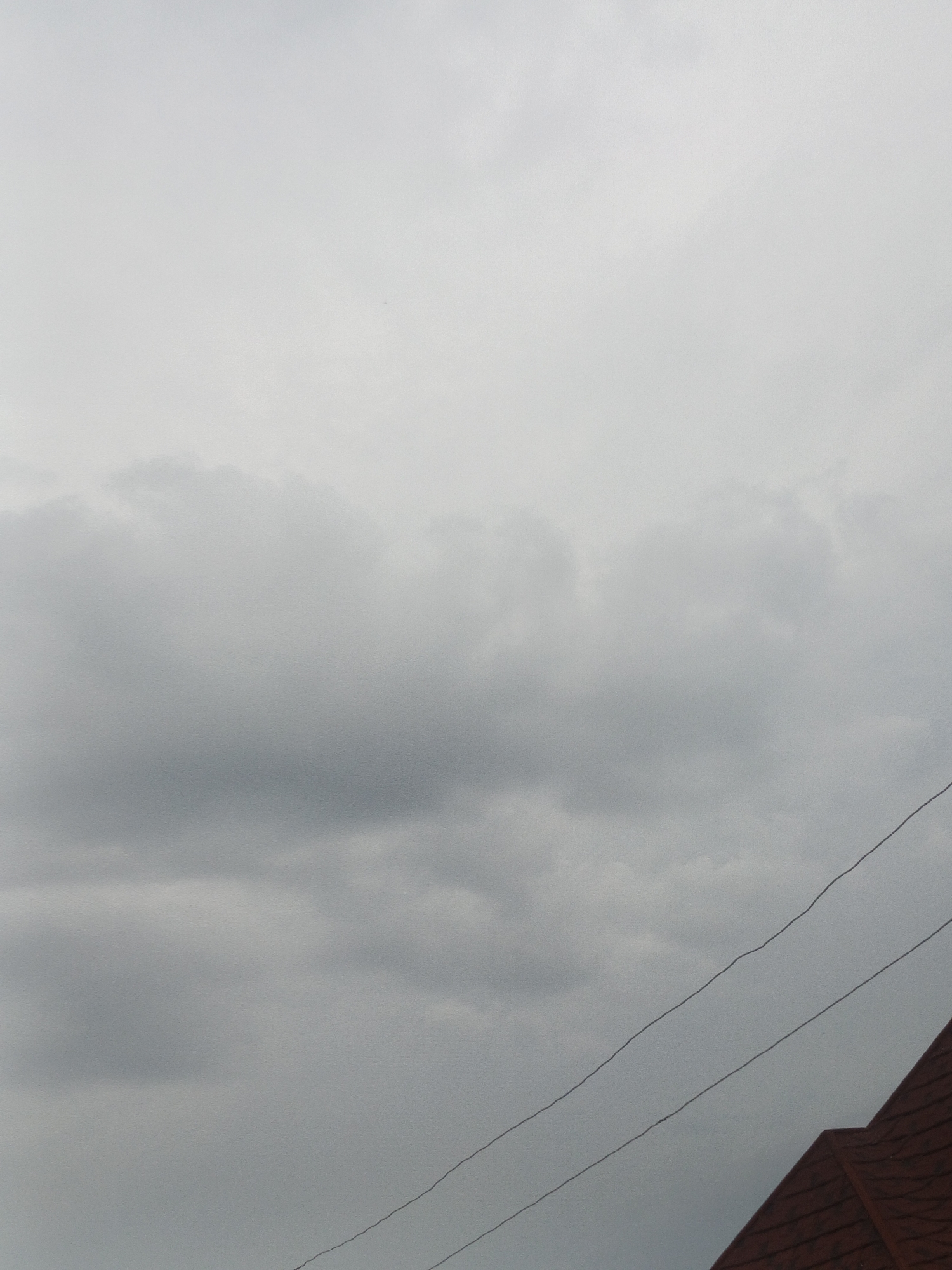 This is a medium shot, eye-level, outdoor photograph taken on a cloudy day, likely in Jos, Nigeria. The majority of the frame is filled with a grey, overcast sky. In the lower right corner, the edge of a reddish-brown tiled roof is visible, angled diagonally upwards. Two thin, dark wires run diagonally across the frame from the lower left to the upper right, crossing over the roofline and into the sky. No people are present in the frame.