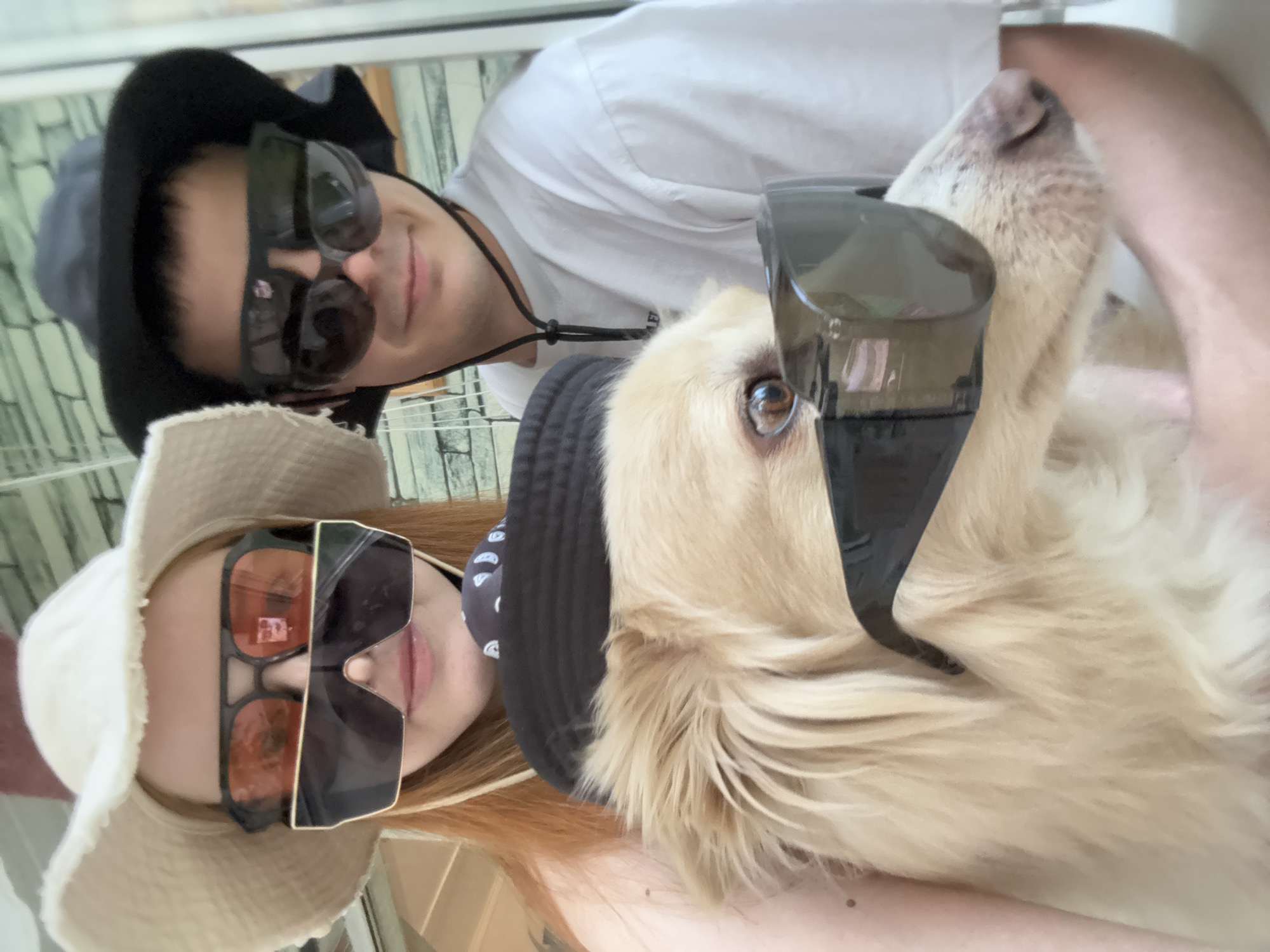 A selfie-style image shows three subjects: a woman with red hair, a man, and a golden retriever dog. The woman is wearing a white sun hat and large, angular sunglasses with orange lenses. The man is wearing a black hat and sunglasses. The dog, positioned centrally, also wears sunglasses and a black hat. The background is indistinct, suggesting an indoor setting. The image is rotated slightly counterclockwise. The location is stated to be Chaikovsky, Russia, though there are no elements in the image to independently confirm the location.