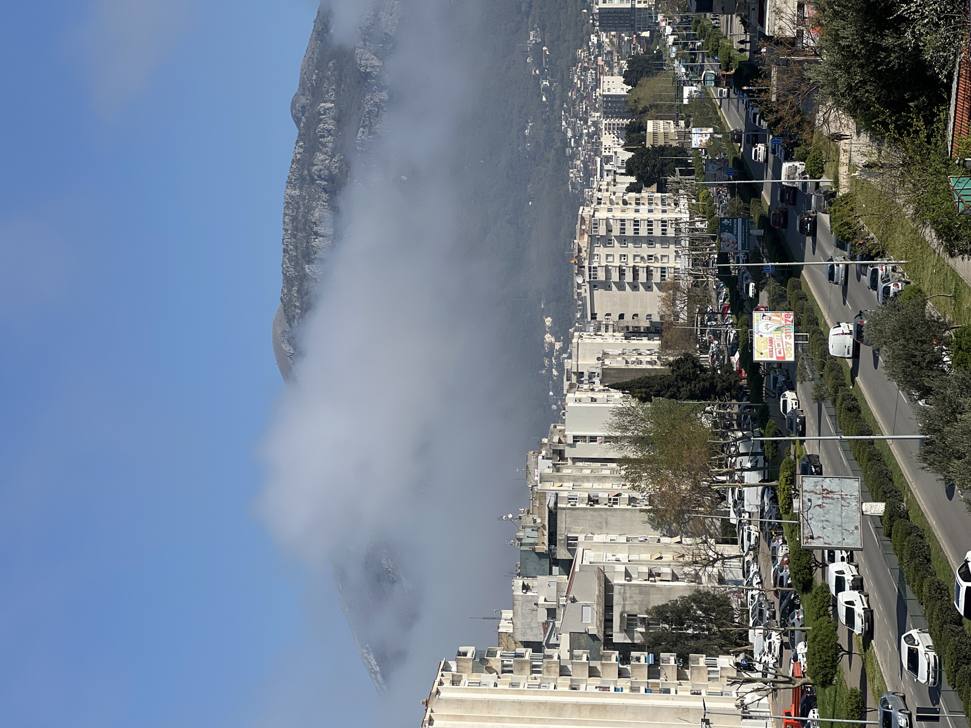 The image depicts a cityscape adjacent to a mountain partially obscured by clouds, likely fog. The location appears to be Bar, Montenegro. The foreground consists of multi-story buildings, characterized by concrete construction and varied architectural styles, possibly residential apartments.
A main thoroughfare runs parallel to the buildings, featuring multiple lanes of traffic with a mix of vehicles including cars, vans, and buses. Streetlights line the median and sidewalk edges. A dense row of trees and greenery separates the sidewalk from the road.
The mountain rises sharply on the left, its rocky terrain partly hidden by a large cloud formation. The sky is clear and blue. There is no visible evidence of pedestrians.