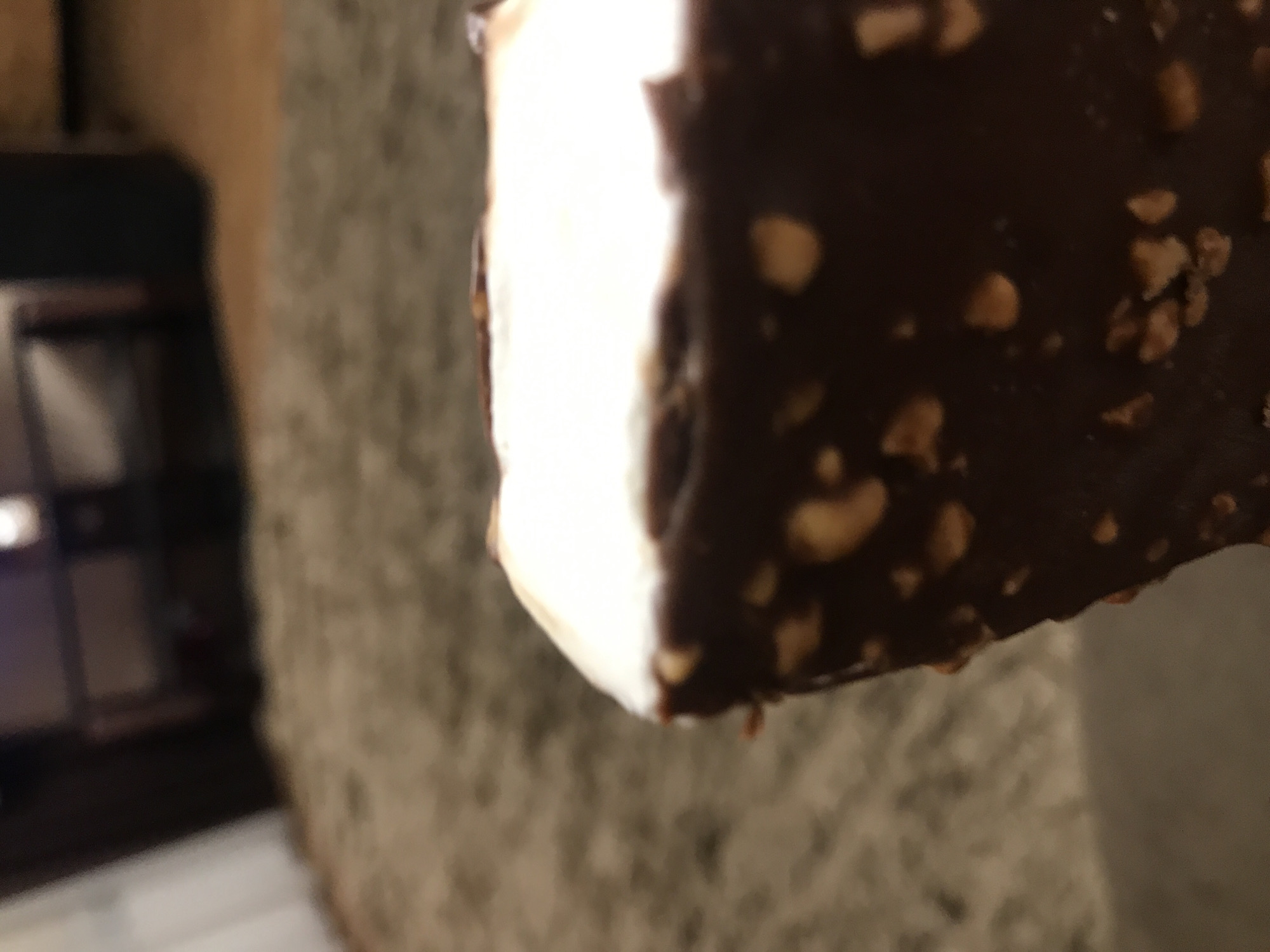 An extreme close-up shot focuses on a partially eaten, square-shaped ice cream bar. The bar consists of white ice cream coated in dark chocolate studded with chopped nuts. The background is blurred, showing a neutral-toned wall with indistinct textures. A dark, blurred object is visible in the lower left corner. The location in Yerevan, Armenia cannot be verified from the image.