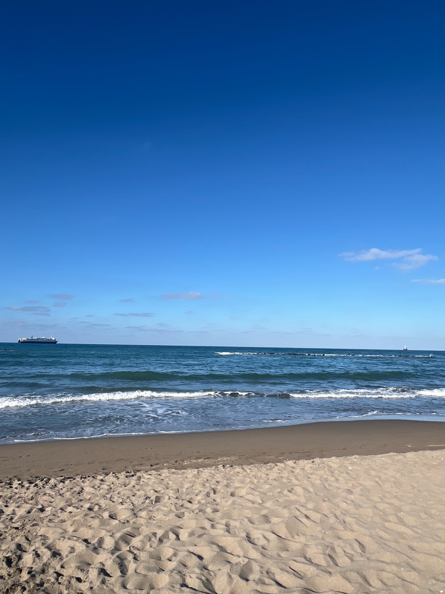The image shows a serene beach scene in Karasu, Turkey. The foreground is dominated by a sandy beach with subtle footprints indicating recent activity. The sand exhibits a light brown hue with variations in texture due to the sunlight. The middle ground features the gentle lapping of waves against the shore, with white foam tracing the edge of the water. The sea itself is a deep blue, stretching towards the horizon.
In the distance, a large cargo ship is visible sailing on the tranquil water, to the left of the frame. Further to the right, a smaller, white structure, possibly a lighthouse or a buoy, marks the horizon. The sky is a vast expanse of clear, bright blue, with a few wispy clouds scattered sparsely across the upper portion of the image. The overall impression is one of peace and natural beauty, characteristic of a coastal landscape.
