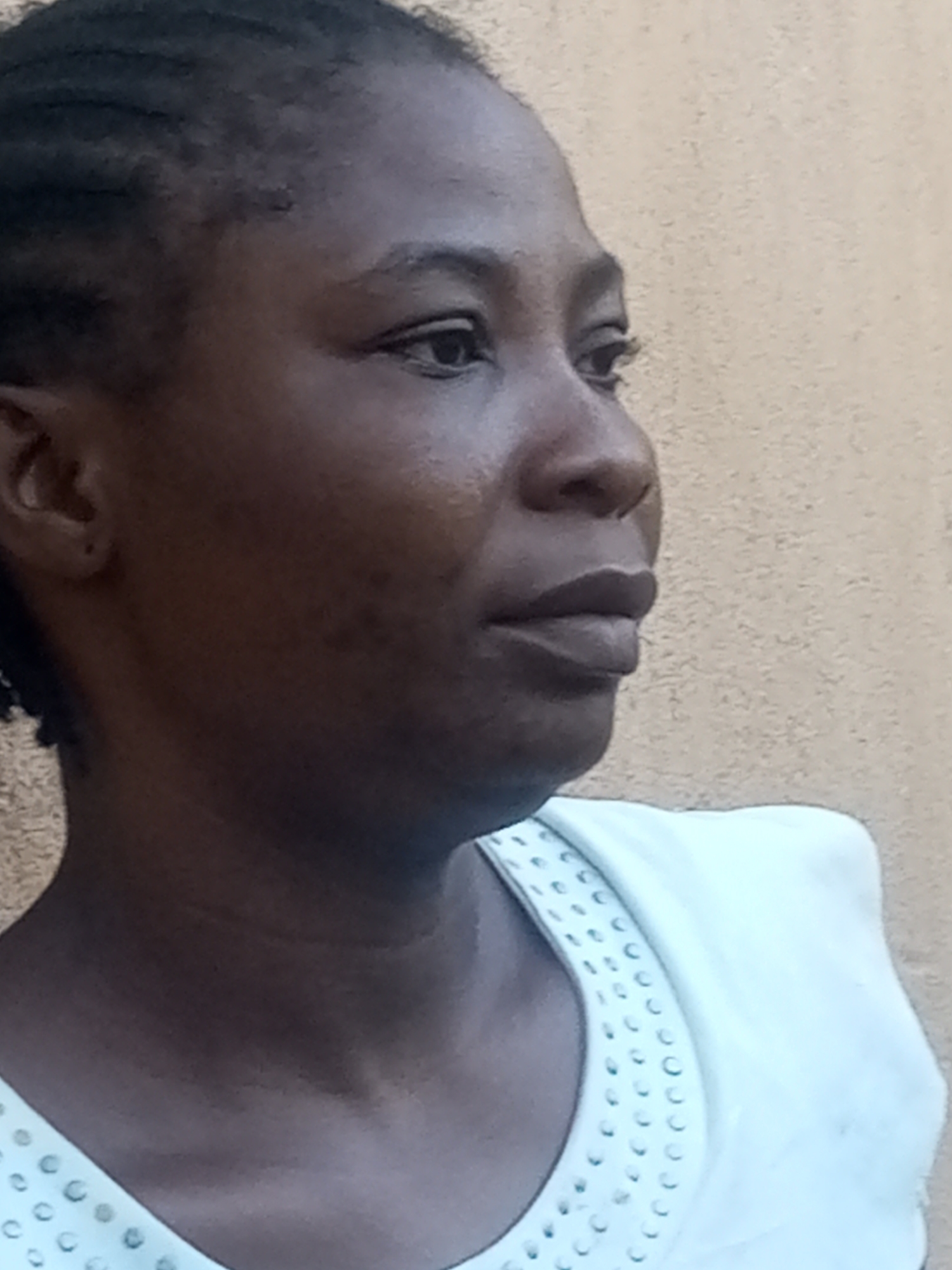 The image is a close-up, profile view of a Black woman, likely an adult, captured outdoors against a neutral, light-colored textured wall. The woman's dark hair is styled in braids that are visible at the top of her head. She is wearing a white garment with a decorative neckline featuring small, circular embellishments. Her skin tone is dark, and her facial features, including her eye, nose, and lips, are visible. The lighting suggests it is daytime. There is no indication of the city of Tempure, Nigeria, within the image itself.