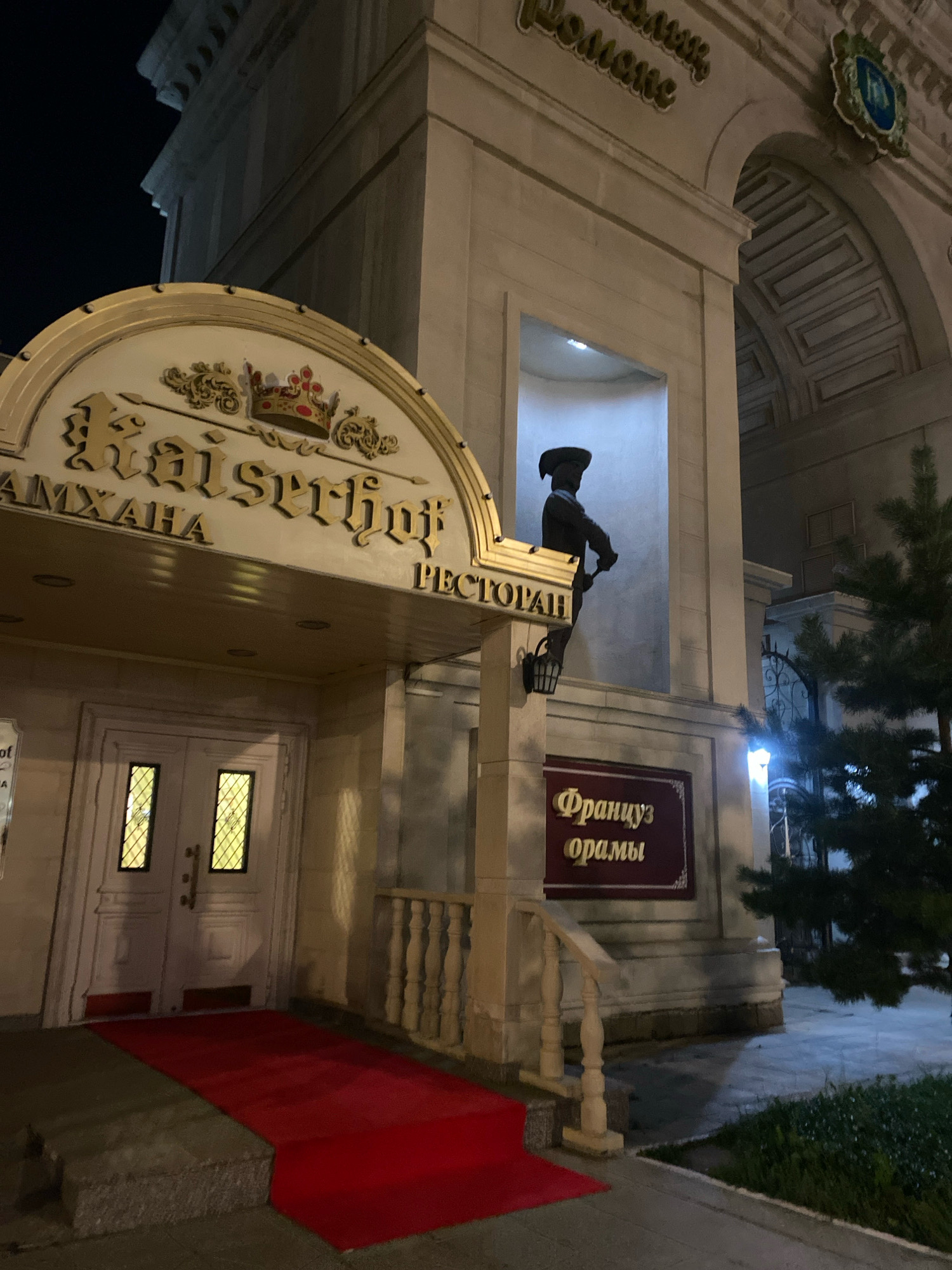 The image shows the entrance to the Kaiserhof restaurant in Astana, Kazakhstan, at night. The restaurant has a large golden sign with a crown and Cyrillic lettering. There is a red carpet leading up to the double doors. To the right of the entrance, a statue of a man in historical dress stands in a niche. Above the entrance, there is another sign with Cyrillic lettering that reads "Француз орамы", which translates to "French street" or "French corner." A large evergreen tree is visible to the right of the building.