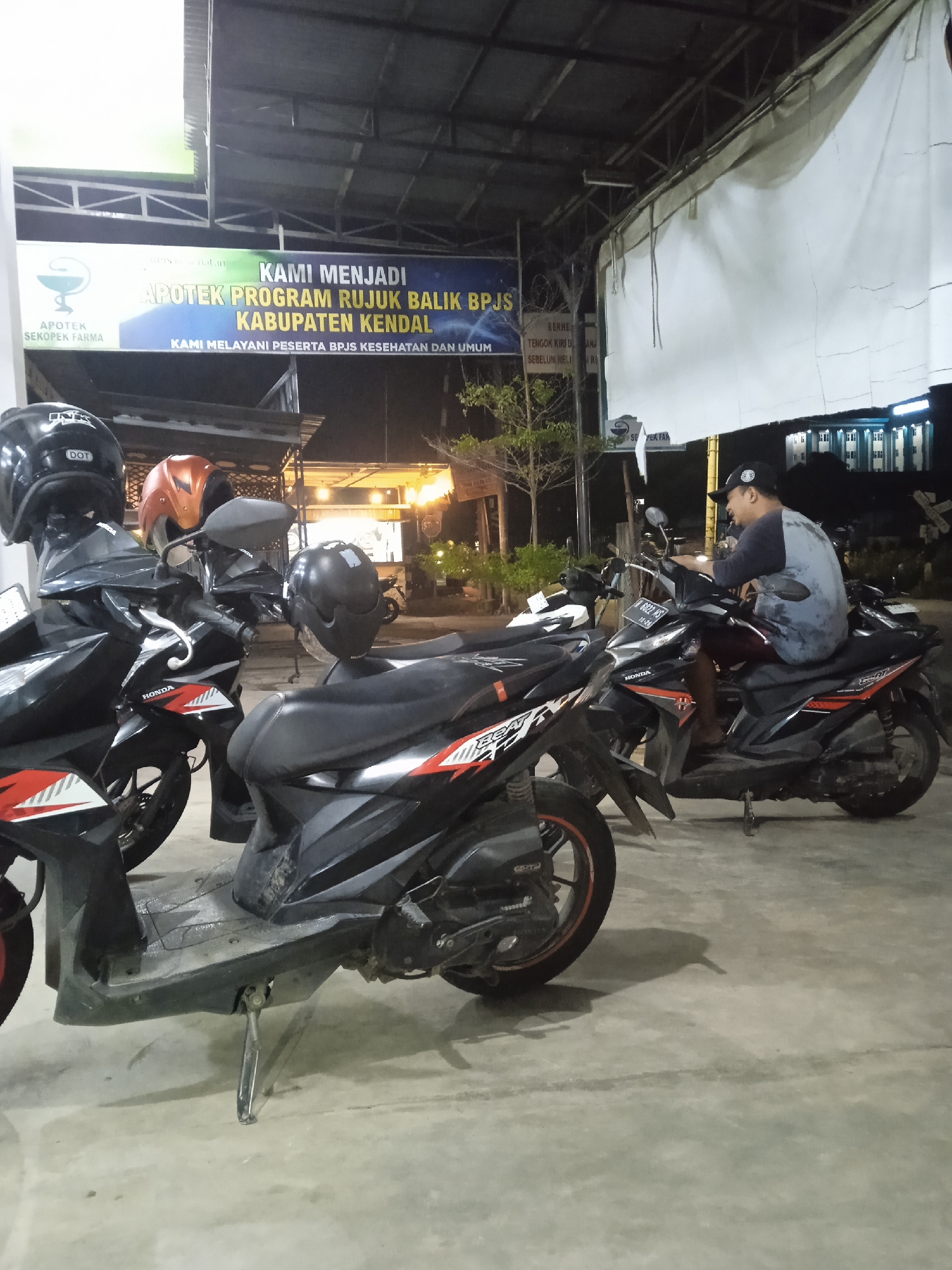 The image captures a nighttime scene in Kebonsari, Indonesia, showing several motorcycles parked on a concrete surface outside a building, likely a pharmacy. The overhead lighting suggests an artificial illumination source.

In the foreground and middle ground, multiple black Honda BeAT scooters are parked, some with helmets resting on their seats or handlebars. One black helmet clearly displays "INK" and "DOT" branding, while another is orange. On the right, a man wearing a dark t-shirt and a baseball cap is seated on one of the black Honda BeAT scooters, with his feet on the ground, suggesting he is either resting or about to depart. The scooter he is on has a visible license plate: "H 6022 MS" with "10-26" below it, indicating a registration for the Kendal region (H is for Central Java, including Kendal) and possibly an expiry date.

Above the motorcycles, a large blue and white banner is displayed, illuminated by an unseen light source. The prominent text on the banner reads: "APOTEK SEKOPEK FARMA," "KAMI MENJADI APOTEK PROGRAM RUJUK BALIK BPJS KABUPATEN KENDAL," and "KAMI MELAYANI PESERTA BPJS KESEHATAN DAN UMUM." This translates to "Sekopek Farma Pharmacy," "WE ARE A BPJS Referral Back Program Pharmacy, KENDAL REGENCY," and "WE SERVE BPJS HEALTH PARTICIPANTS AND THE GENERAL PUBLIC." Another, less clear sign is partially visible to the right of the banner, containing phrases like "BERNE," "TENGOK KIRI DE ANA," and "SEBELUM MELI R," which seem to be warning or instructional signs. The background is dark, with some distant lights from other buildings visible.