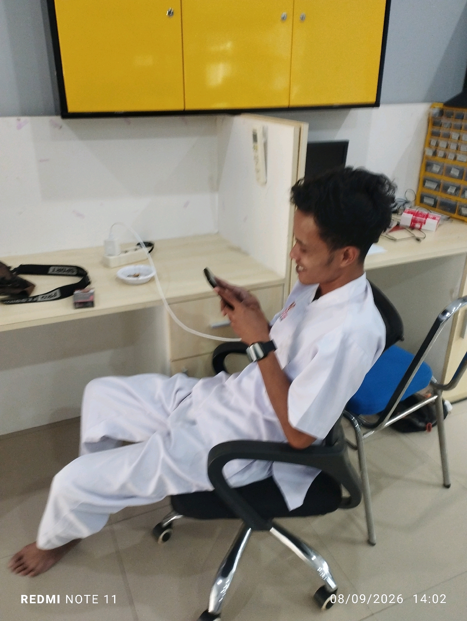 A young man in a white uniform sits on a black office chair with a blue cushion, looking at his phone. He is in a room with a desk, yellow cabinets, and a grid of small drawers. A camera strap and a pack of cigarettes are on the desk. The man's right foot is bare. The image is dated 08/09/2026 14:02 and includes the text "REDMI NOTE 11".