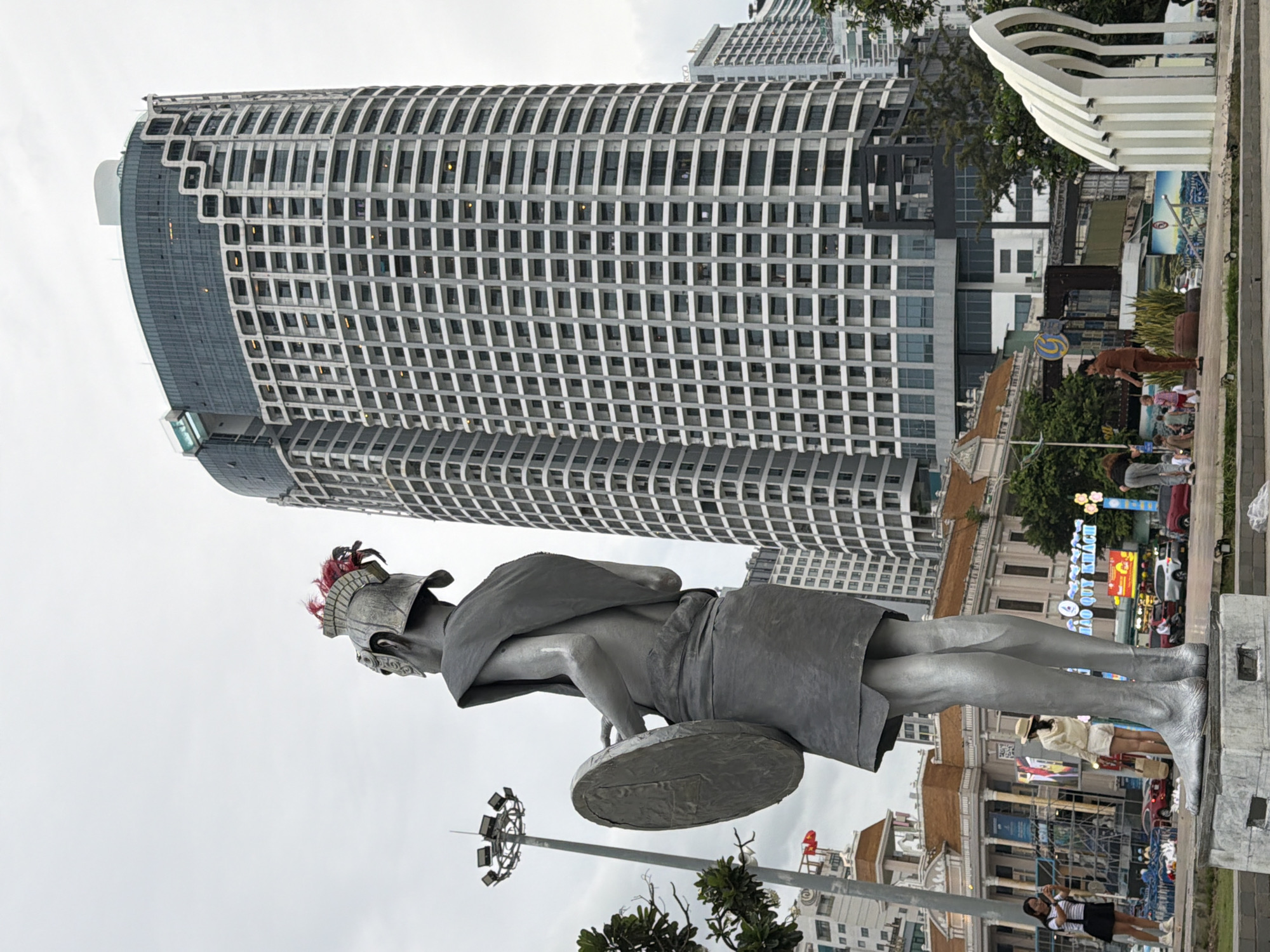 A full-shot, eye-level exterior photograph taken on an overcast day in front of the Vinpearl Luxury Hotel in Nha Trang, Vietnam, features a monumental silver statue of a warrior adorned with a feathered helmet, holding a round shield and a sword, with its back to the viewer. The statue stands on a stone pedestal, its metallic surface reflecting the dull sky. In the background, the curved, modern facade of the Vinpearl Luxury Hotel, with its grid of windows, dominates the skyline. To the left of the hotel, a tall, circular light pole with multiple spotlights stands prominently. Below the statue, a paved area is visible with several people walking and observing. To the right, a series of abstract white sculptures creates a rhythmic, arched pattern. The overall scene depicts a public space with prominent architectural landmarks and urban elements.