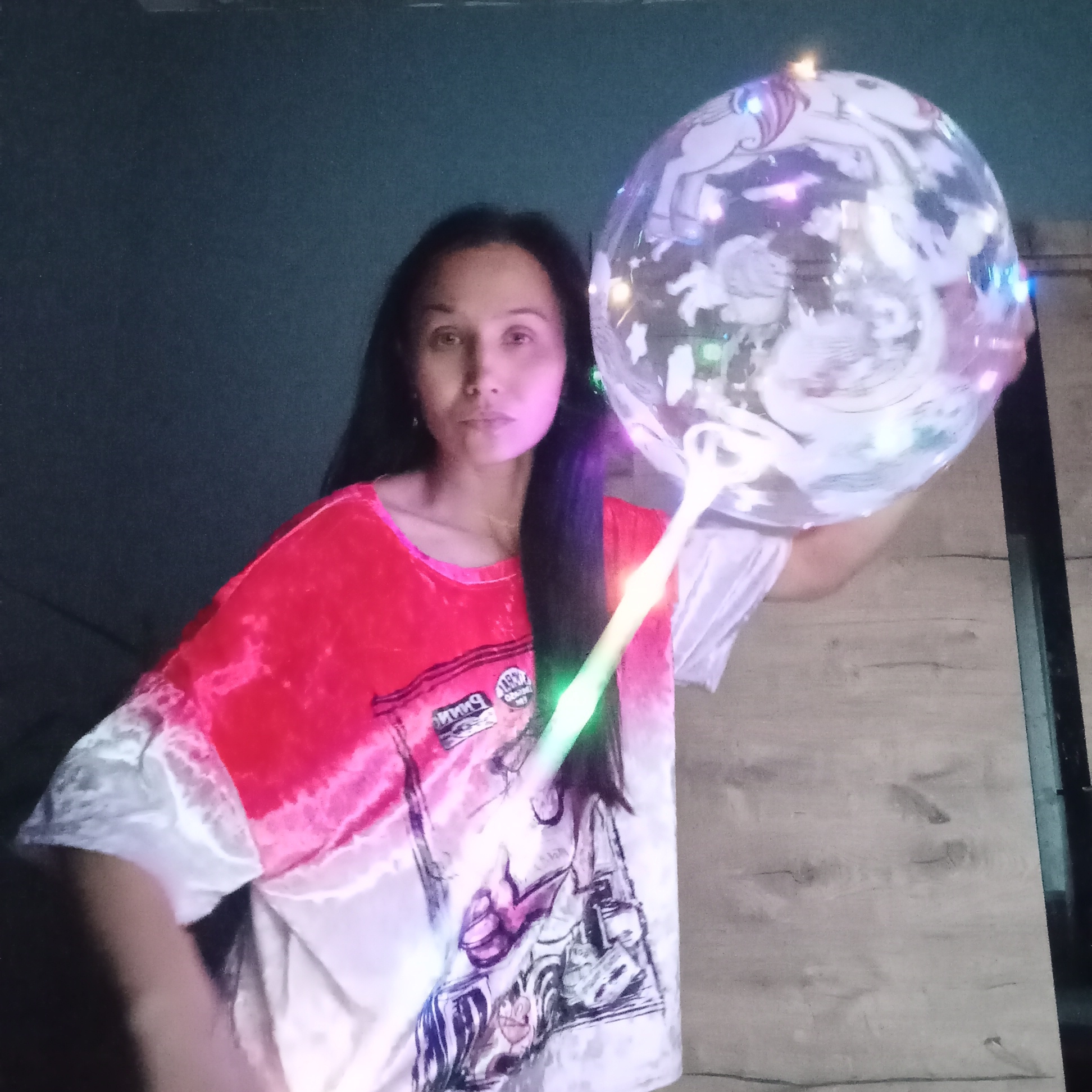 An individual is depicted indoors, positioned slightly right of the frame's center. The subject is a woman with long, dark hair, looking directly at the viewer. Her face is partially illuminated by a magenta glow. She wears a short-sleeved t-shirt featuring a red upper section with a tie-dye transition to white on the lower half and sleeves. Graphics are visible on the front of the shirt, including text that appears to be "PINK" in reverse print.
With her right hand, she holds a transparent spherical balloon attached to a white stick. Both the balloon and the stick are internally illuminated with multi-colored LED lights, casting purple, pink, blue, and green hues. The balloon features white-outlined illustrations of unicorns. The background consists of a dark, uniform surface on the left and upper portions, and a light-colored wooden panel surface on the right, possibly part of furniture or flooring. The overall lighting is dim, with the primary light sources emanating from the held objects. The scene is set indoors in the city of Pervomaysky, Russia.