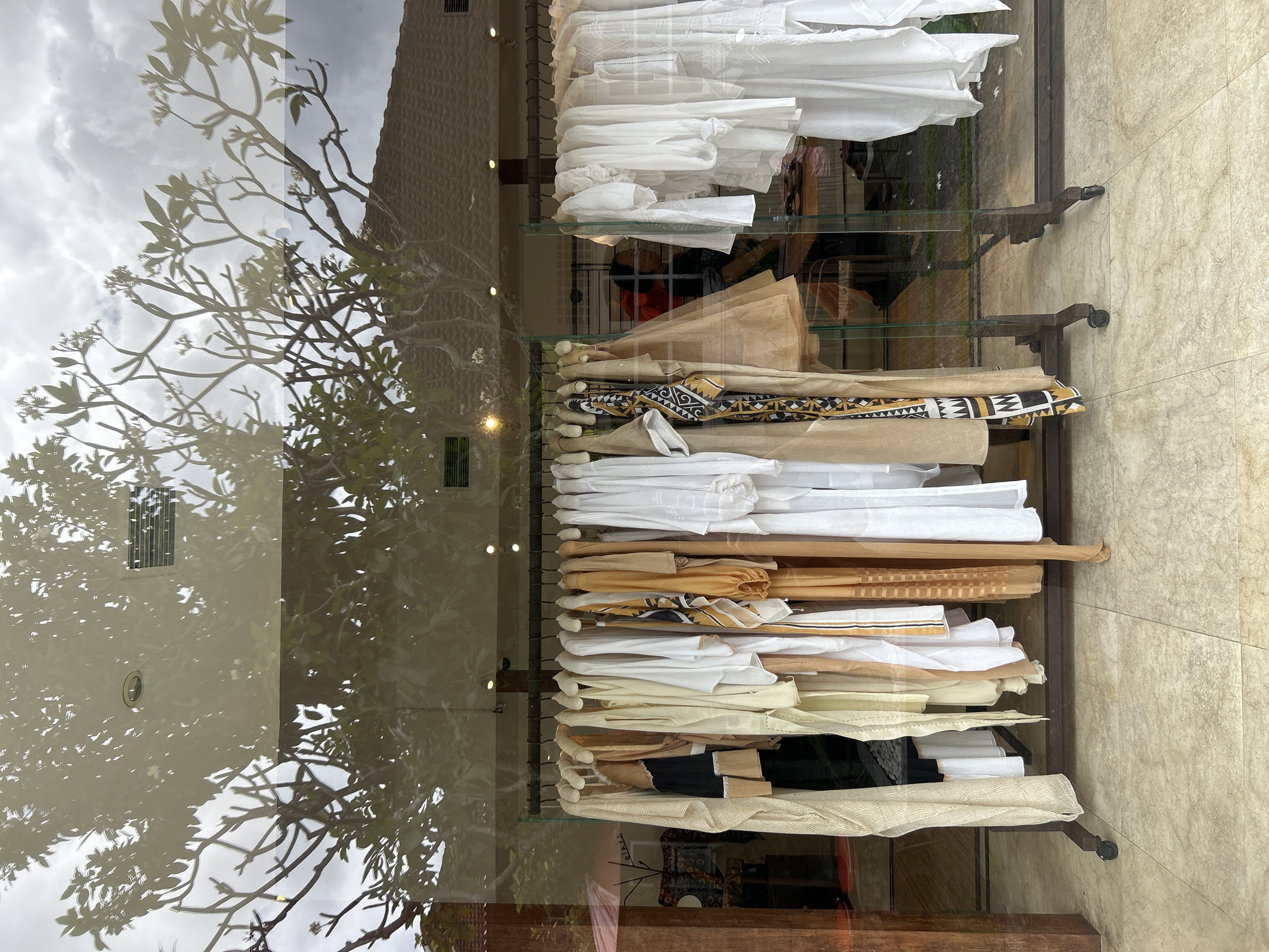 This is a medium shot, eye-level photograph taken outdoors in front of a store in North Kuta, Indonesia. The scene is dominated by a reflection of a tree and cloudy sky on a store window. Inside the store, a rack of clothing is displayed. The clothing consists of several long, flowing garments in light colors, primarily white, beige, and tan. One garment on the left is a long, cream-colored sheer robe. Another garment towards the center features a black, white, and yellow geometric pattern. Behind the clothing rack, the interior of the store is visible, with shelves and a person wearing a red top. The floor outside the store is tiled with a marble-like pattern. The overall lighting is soft and diffused, suggesting an overcast day.