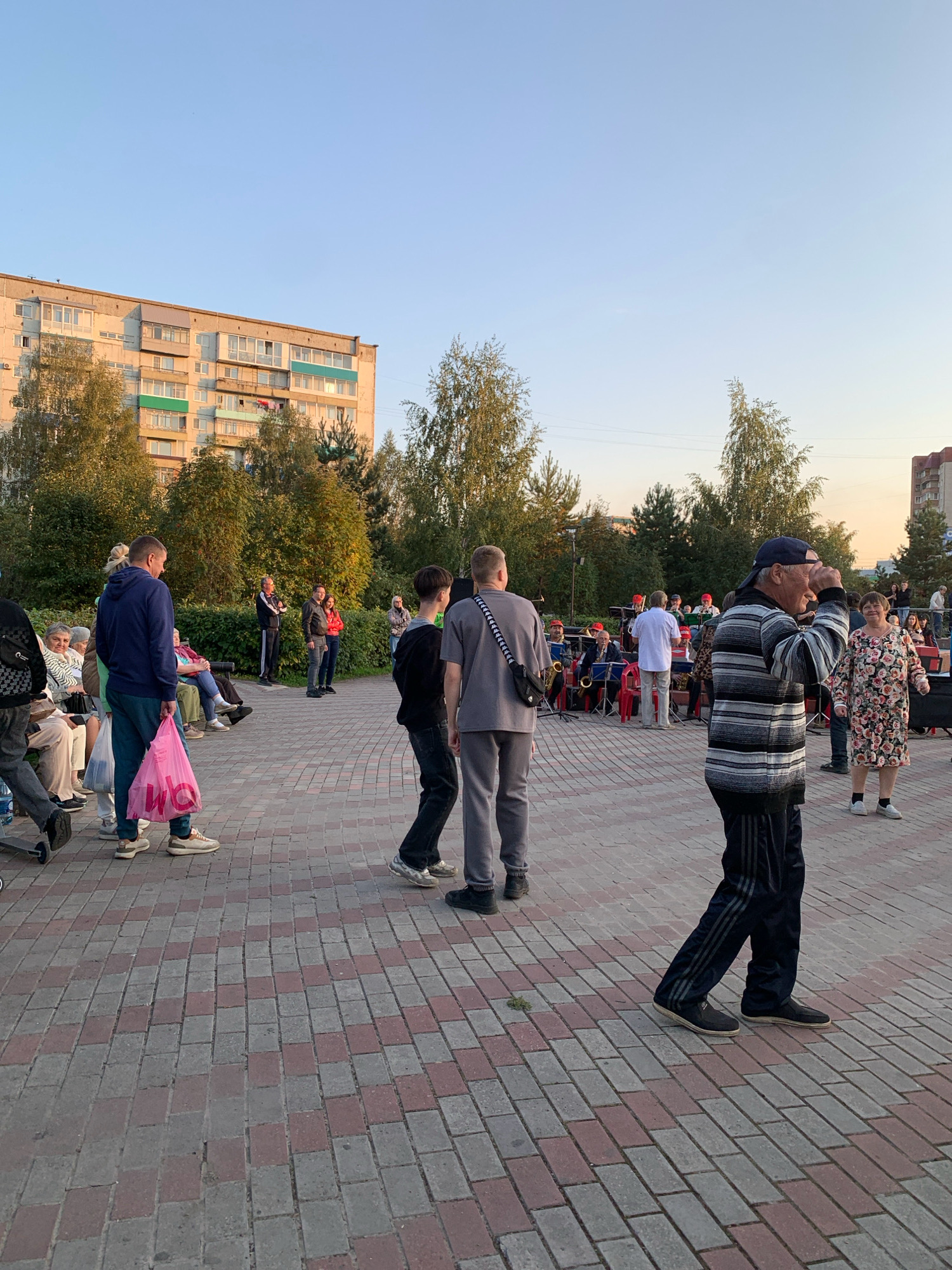In Prokopyevsk, Russia, a crowd gathers in an outdoor public space, possibly a park or square, paved with red and grey interlocking bricks. To the left, a multi-story residential building, typical of Soviet-era architecture, stands against a clear blue sky. Trees with autumn foliage surround the area. A band with brass instruments is set up in the background, with musicians in uniform red hats and white shirts. Spectators are seated on benches and standing around. In the foreground, several individuals are captured in motion. A man in a striped sweater and a baseball cap is looking towards the right and appears to be shielding his eyes from the sun. To his right, a woman in a floral dress smiles. To the left of the man in the striped sweater, a younger man in a grey sweatshirt and dark pants walks away from the camera. Further to the left, another man in a blue hooded sweatshirt carries a pink shopping bag. Other individuals are visible in the midground, some seated on a bench. The overall scene suggests a community event or performance taking place during the late afternoon.