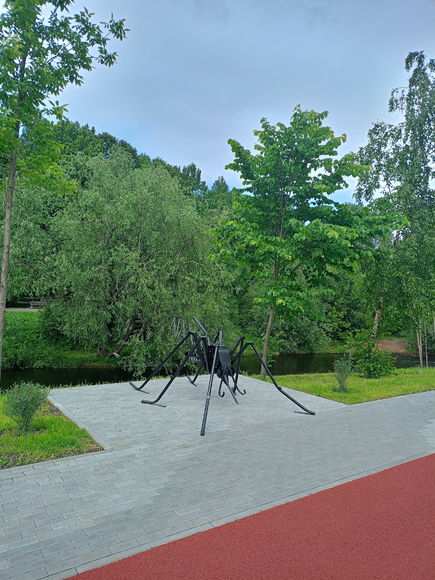 A black metal sculpture resembling a large spider stands on a grey brick pathway in a park. The spider sculpture is the main subject, with its intricate metal legs spread out. The pathway is bordered by green grass and a dark body of water, likely a river or pond, which is surrounded by lush green trees and foliage. In the background, more trees and vegetation create a dense, natural landscape. The sky above is overcast with a light grey hue. The scene appears to be set during the daytime, with diffused light suggesting cloudy weather. The overall atmosphere is peaceful and natural, with the sculpture adding an element of artistic interest to the park setting.