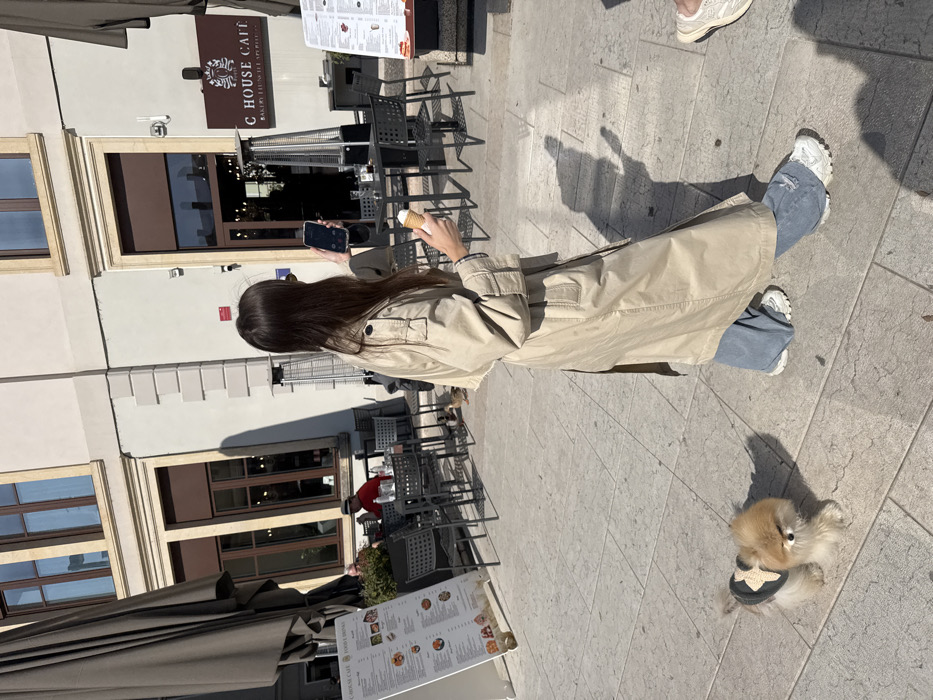 The scene depicts a sunny outdoor cafe area in Piazza Ferdinando di Savoia, Peschiera del Garda, Italy. A female subject, with long dark hair, is walking from left to right across a paved pedestrian area. She wears a beige trench coat, blue wide-leg jeans, and white sneakers. Her left hand holds a dark-colored smartphone, positioned as if taking a photograph or video. Her right hand holds an ice cream cone with white ice cream. Ahead and slightly to her left, a small, fluffy light-brown dog, possibly a Pomeranian, walks on a leash not visible, wearing a dark vest with a light-colored bow-shaped embellishment.
To the right of the subjects, the outdoor seating of "C HOUSE CAFE" is visible, featuring multiple black metal tables and chairs. Two tall, silver outdoor heaters stand among the tables. A brown sign on the building above the cafe entrance reads "C HOUSE CAFE BAKERY BRUNCH APERITIVO". The building features light-colored walls with prominent window and door frames in a dark brown hue. To the far left, a large, dark brown umbrella extends over additional cafe seating. Two freestanding menu boards, one displaying "C HOUSE CAFE FOOD & DRINKS", are positioned near the cafe tables. A man in a red shirt and a hat is seated at one of the tables in the background. Several small ducks are present on the pavement beneath the cafe tables. The ground is paved with light-colored rectangular tiles, and strong shadows indicate direct sunlight.