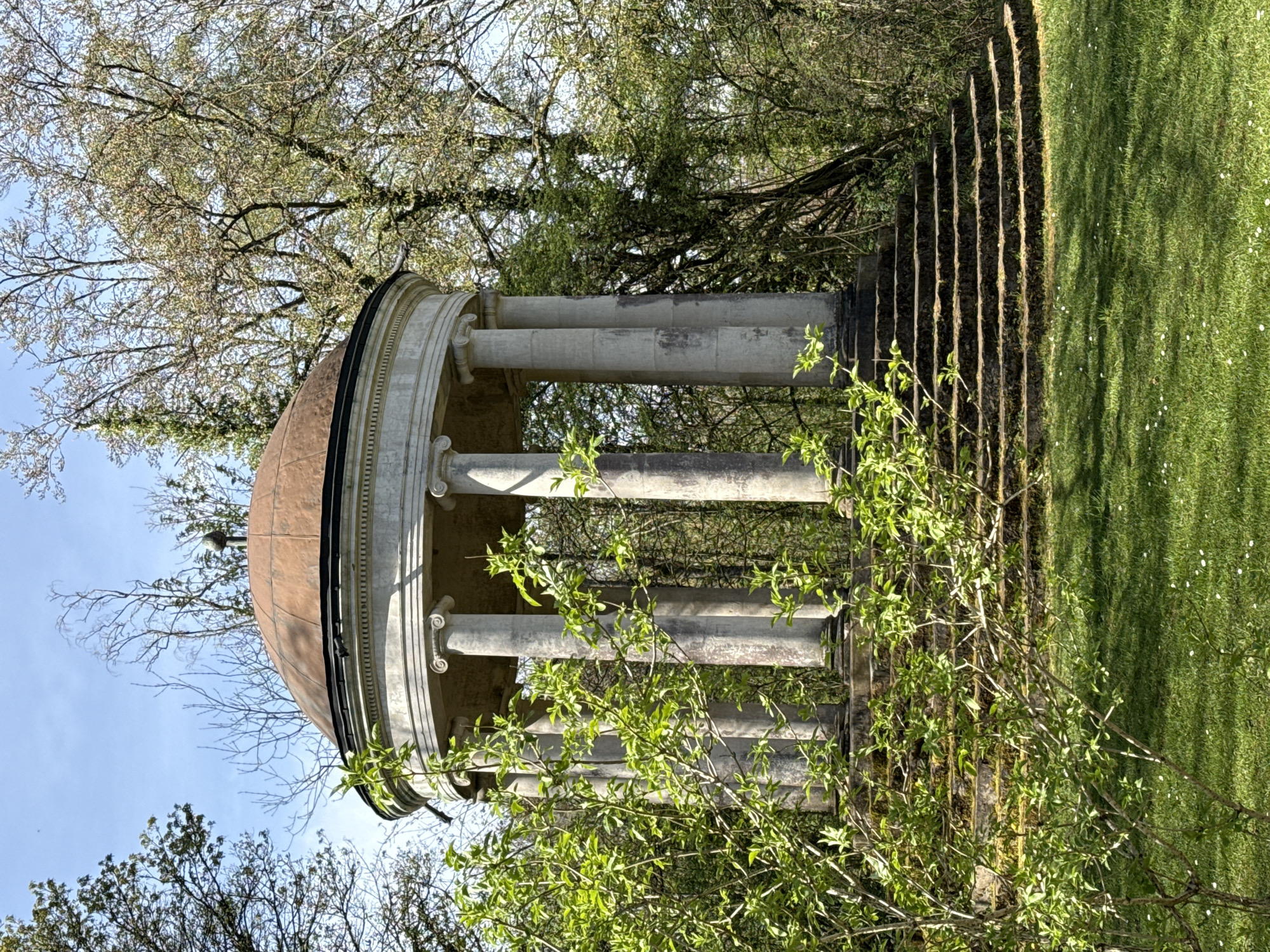 A stone gazebo with a dome-shaped roof stands in a grassy area. The structure is supported by multiple pillars. A set of stairs leads up to the structure. Trees surround the gazebo. The location is likely Esch-sur-Alzette, Luxembourg.