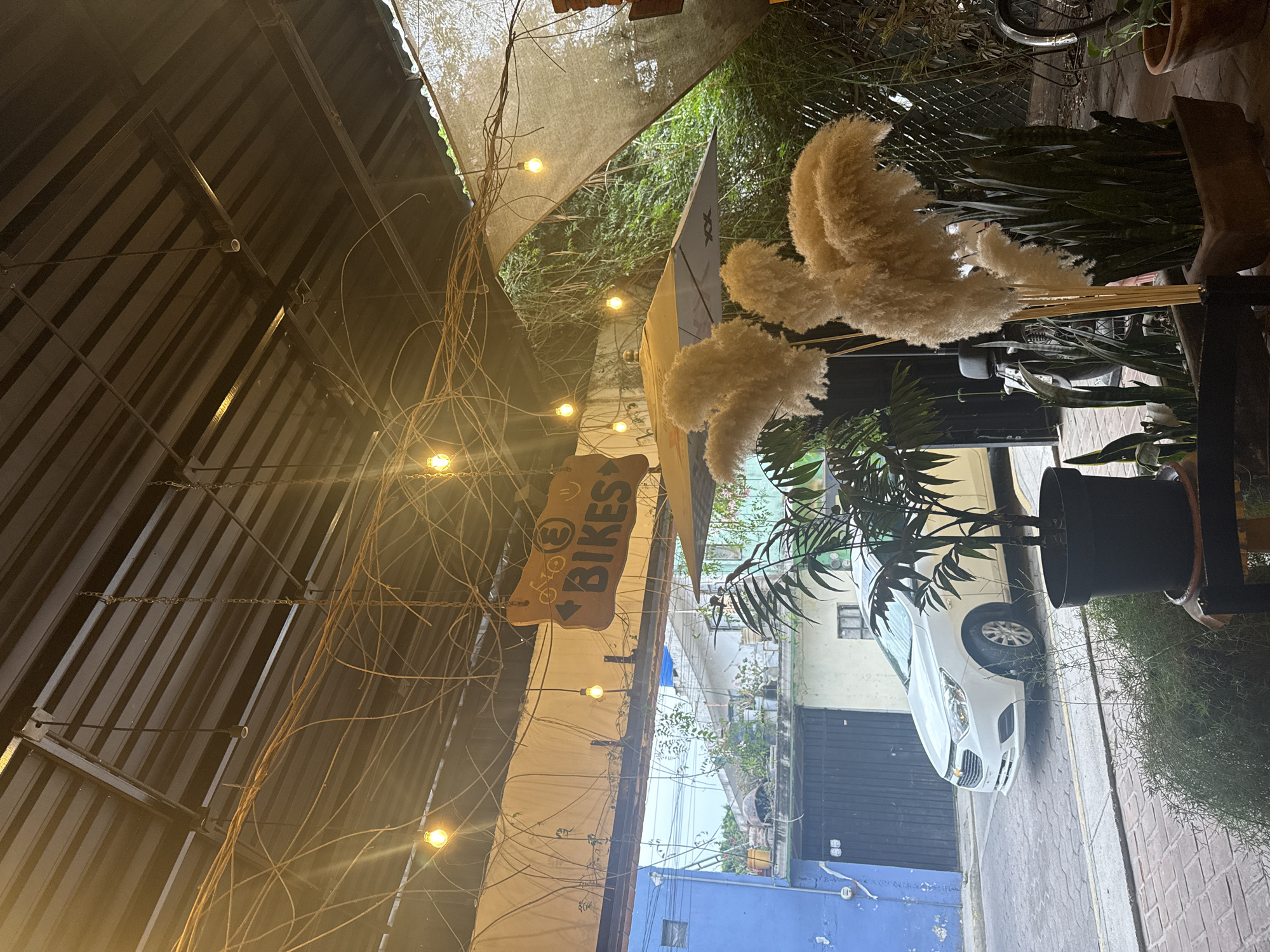 The image depicts an outdoor space in San Andrés Cholula, Mexico, likely a patio or terrace. The space is partially covered by a metal roof with visible beams and string lights, creating a dim, warm ambiance. Decorative elements include dried branches and pampas grass, adding texture and visual interest. A wooden sign with "BIKES" is visible. Beyond the partially covered area, a street scene unfolds. A white car is parked along the curb of a narrow street, flanked by buildings with varying architectural styles, including a blue building and buildings with visible windows and garage doors. The sky is overcast, suggesting a subdued light.