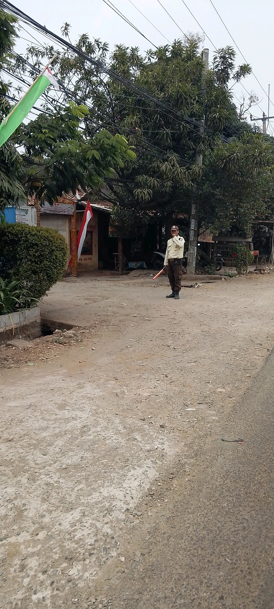 A man in a light-colored uniform and dark pants stands in a dirt road in Cileungsi, Indonesia, holding a red signaling baton. He is wearing a cap and appears to be gesturing with his right hand. The background features trees, power lines, and a small building with a red and white flag hanging outside. A green bush is visible on the left side of the image. The road is unpaved and has some debris scattered on it.