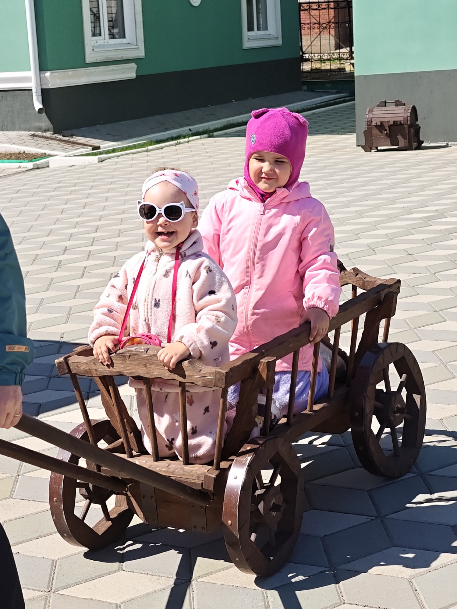 Two young girls are in a wooden cart being pulled by a person whose arm and hand are partially visible on the left side of the image. The girl in the front is wearing a pink outfit with a rabbit pattern, a pink headband, and white cat-eye sunglasses. She is smiling. The girl behind her is wearing a pink jacket and a pink hat. The cart has four wooden wheels.
The scene is set on a paved area of interlocking gray tiles. In the background is a green building with white trim, a fence, and a wooden chest. The image appears to be taken in daylight.
The location is likely Chaikovsky, Russia, given the prompt's reference to this city. However, there are no readily apparent landmarks that would confirm the exact location within the city.