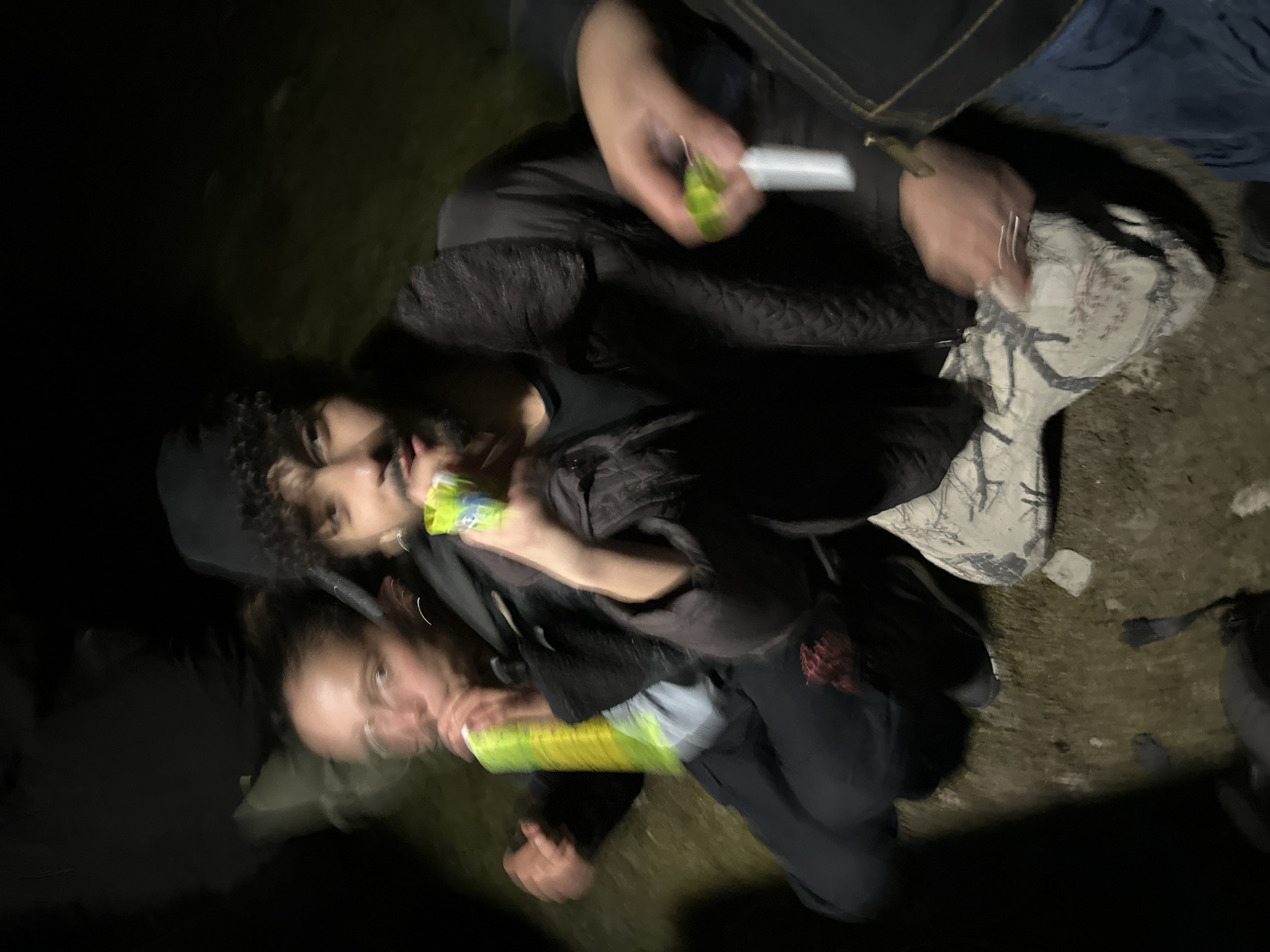 In a dark outdoor environment, three people are seen. One individual in a black hat and jacket holds a yellow object, while a second person beside them appears to be holding a similar item. A third individual, wearing a dark jacket and jeans, holds an object in their hand. The surroundings consist of ground cover and possibly a grassy area. The location is presumed to be Auvers-Saint-Georges, France.