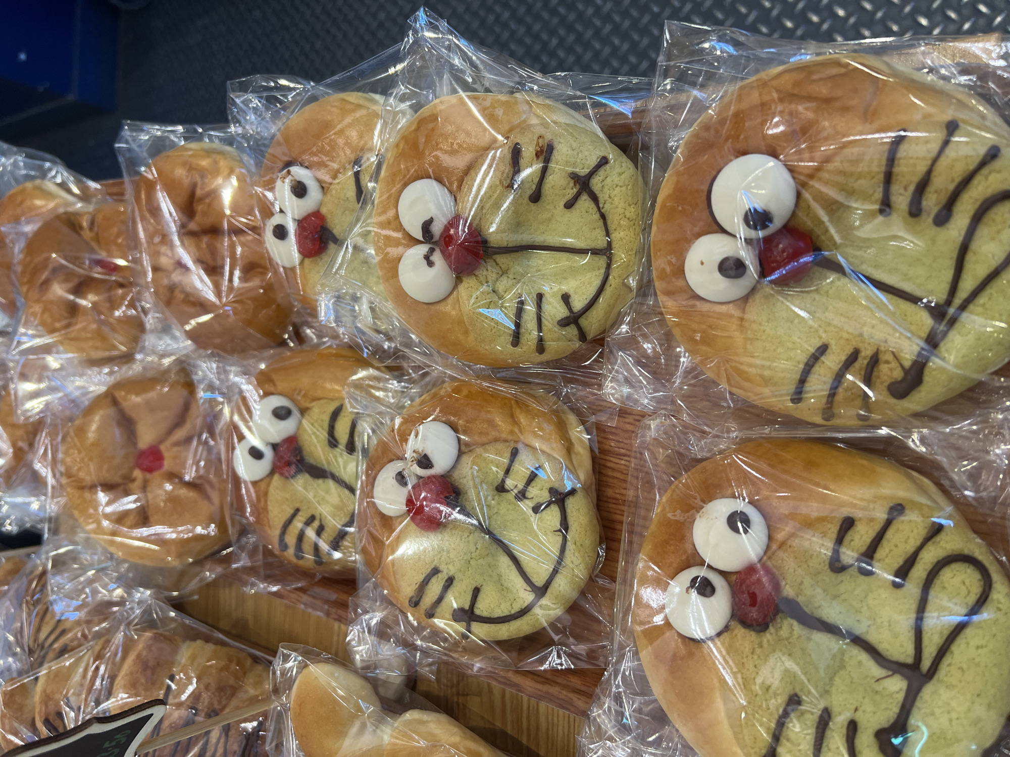 The image displays a retail display of individually plastic-wrapped baked goods on a light brown wooden surface. The central focus is on eight round buns, each decorated to resemble the face of the character Doraemon. These buns feature a yellowish-green flat top, a red spherical nose, two prominent white circular eyes with black pupils, and a smiling mouth with whiskers drawn in dark chocolate. They are arranged in rows, with four buns clearly visible in the foreground and others receding.
To the upper left, partially visible, are other plastic-wrapped pastries, including a plain golden-brown bun and a croissant-like item with dark stripes. A small black sign with white lettering is visible in the extreme upper left corner. The wooden surface extends across the lower and central areas, while a dark, textured surface, possibly a metal floor plate or shelving, is visible on the right side and in the background.
The scene depicts a commercial setting, likely a bakery or grocery store, presenting specialty character buns for sale. The location is in New York, United States of America. The precise street address or specific neighborhood within New York City cannot be determined from the visual evidence presented. No individuals, actions, or direct interactions are visible.