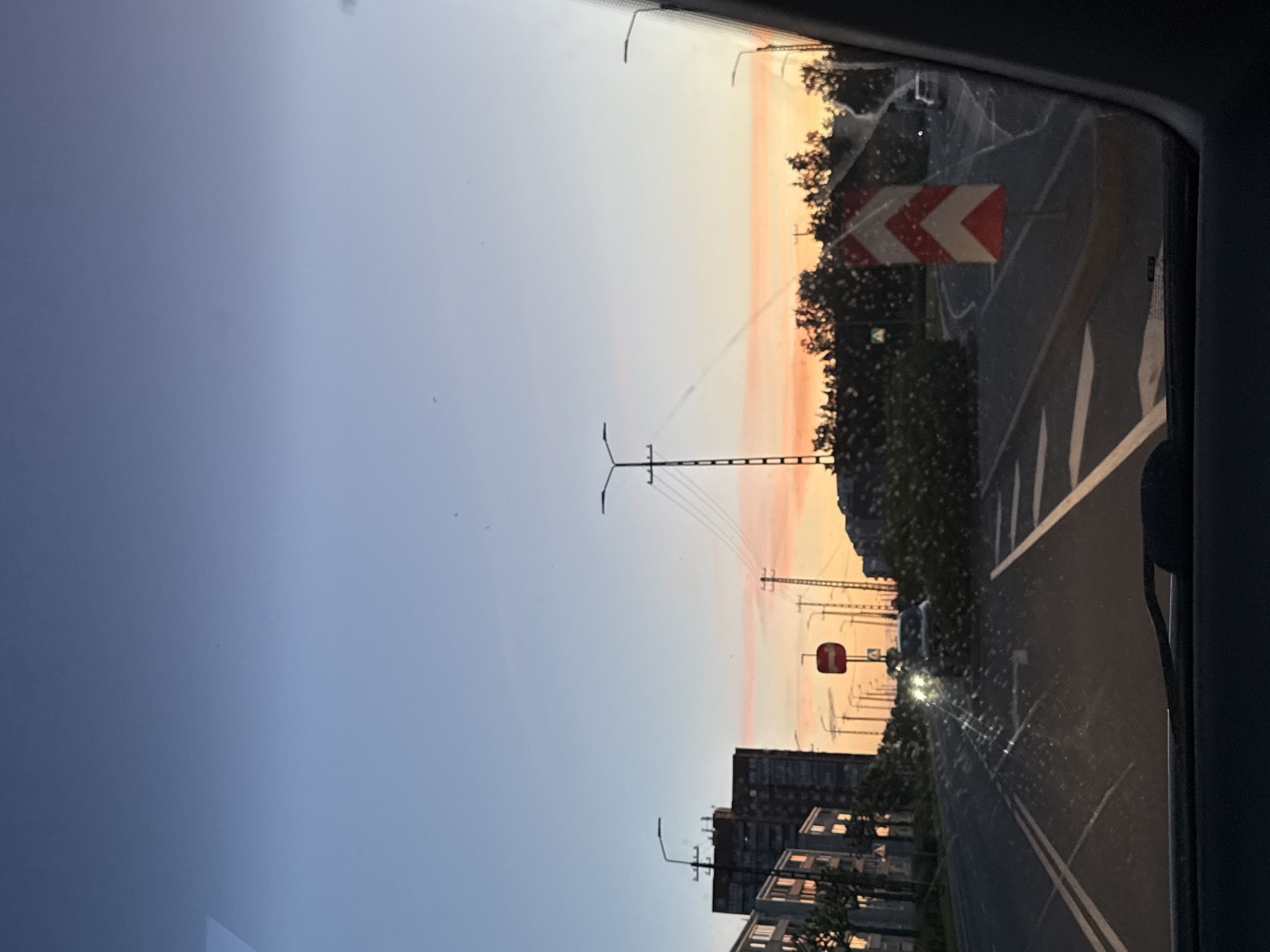 The image, taken from inside a vehicle, depicts a city street in Tallinn, Estonia, during what appears to be either sunrise or sunset. The sky displays a gradient of colors, transitioning from a clear blue at the top to soft oranges and pinks near the horizon.
On the left side of the frame, a row of apartment buildings, characterized by multiple floors and windows, lines the street. Streetlights with modern designs are visible along the road, some with visible electrical wires running between them. In the distance, a traffic sign with a red circle and a white horizontal bar is mounted on a pole, indicating a prohibition.
The road itself is asphalt, marked with white lines. A car with its headlights on is visible in the middle of the road, moving away from the viewer. On the right side of the road, a prominent traffic sign with red and white chevron markings directs traffic to turn. Trees and foliage are present along the sides of the road, with some appearing to be overgrown or recently affected by weather. The windshield of the vehicle shows some smudges and reflections. The overall impression is of an urban environment experiencing a transition in natural light.