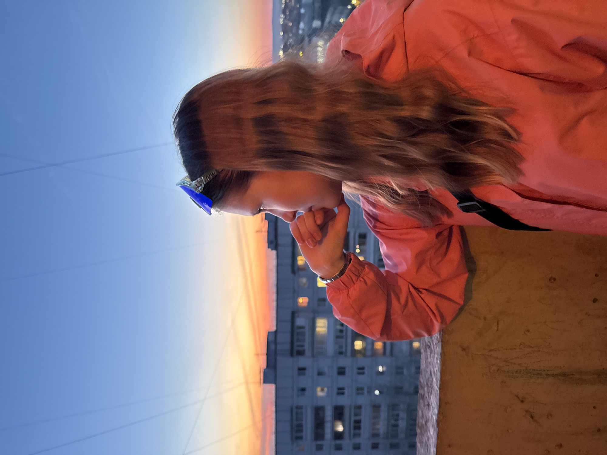 A woman in a pink jacket stands on a balcony overlooking a cityscape, possibly in Chaikovsky, Russia. She rests her chin on her hand and appears contemplative. The sky behind her is a gradient of blue and orange, suggesting either sunrise or sunset. A tall apartment building is visible below, its windows lit. She is wearing blue sunglasses.