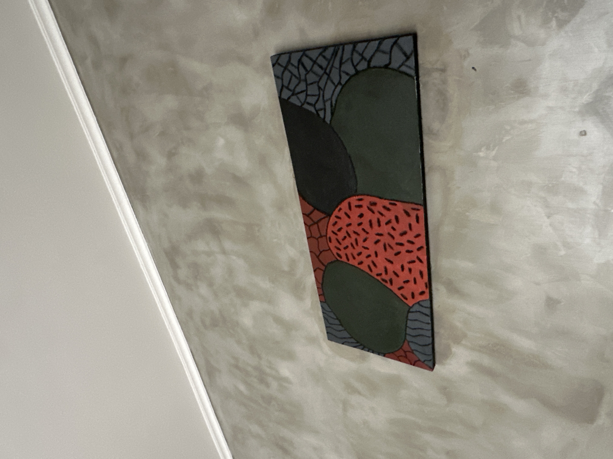 A rectangular, abstract painting hangs on a textured gray wall in North Kuta, Indonesia. The painting is horizontally oriented and features a design of irregular, overlapping shapes in shades of red, dark green, black, and gray. One shape, primarily red, is filled with small, dark speckles. Another gray shape is outlined with a pattern of curved lines. The wall has a rough, concrete-like finish. A white crown molding runs along the top of the wall, meeting a white ceiling. The lighting appears to be natural, casting subtle shadows and highlighting the texture of the wall.