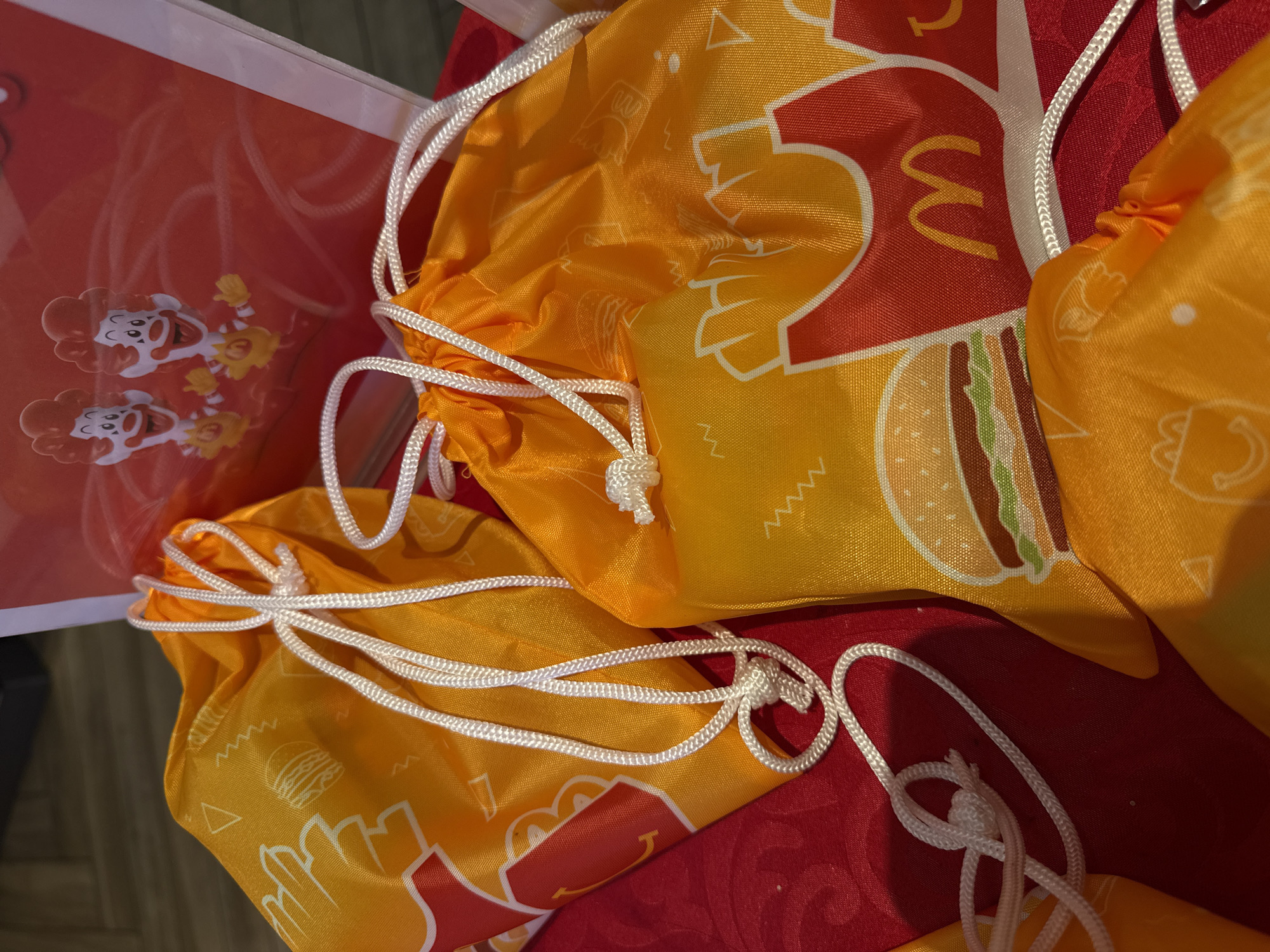 A close-up, eye-level, outdoor shot shows an assortment of bright orange drawstring bags with a pattern of french fries and hamburgers, and the McDonald's logo. The bags are arranged on a red, textured surface, possibly a tablecloth. In the background, a red poster featuring two cartoon clown figures and the McDonald's logo is partially visible, along with a glimpse of wooden flooring. The scene appears to be a promotional display, possibly in a commercial setting. There are no people or discernible human activity present in the frame. The lighting suggests it is daytime, with a slight glare on the fabric of the bags. The precise location in Nanshan District, China, cannot be determined from the visual information provided.