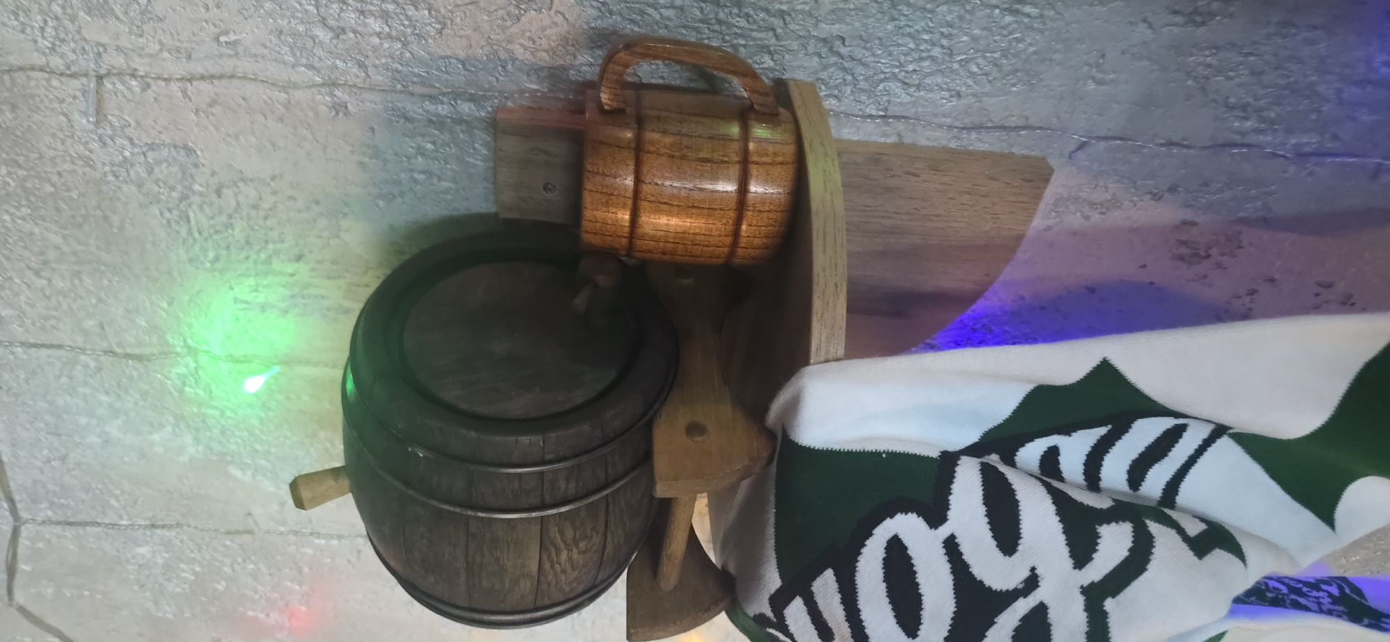 This is a close-up, vertical, eye-level shot of a wooden barrel and a wooden mug on a wooden shelf, possibly in an apartment in Krasnodar, Russia. The shelf is mounted on a textured, light-colored wall, which appears to be a concrete or plaster surface with an uneven finish.
To the left, a dark wooden barrel with metal hoops is positioned horizontally. A small wooden spigot protrudes from the right side of the barrel. The barrel is supported by a wooden rack with two angled legs. Hanging over the front of the shelf, partially obscuring the rack and the lower portion of the barrel, is a white and green knitted fabric, likely a scarf or blanket, with a repeating pattern of black and white elements.
To the right of the barrel, on the same shelf, is a brown wooden mug with a handle. The mug has a banded design around its body. Behind the mug, a small, light-colored wooden block is mounted to the wall.
The scene is illuminated by a string of lights, evident by the blurry green and red light spots on the wall behind the barrel. There is also a purple light source visible from below, casting a glow on the lower part of the shelf and the wall. No people are present in the image, and no specific actions are depicted. The overall impression is of a decorative display, possibly in a home setting.