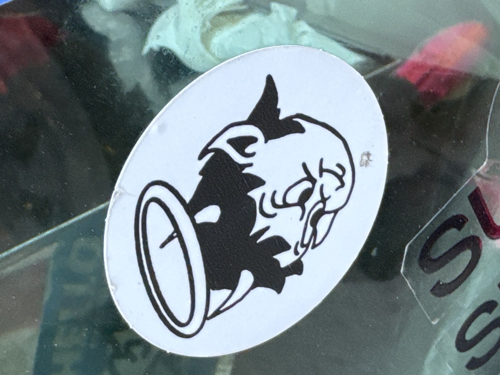 An oval sticker with a black-and-white design depicting a boar's head with a halo is affixed to glass or a similar transparent surface. Adjacent to the sticker is a partial view of another sticker featuring the letter "S" in black, suggesting multiple stickers are present. The photograph was taken in Milan, Italy.