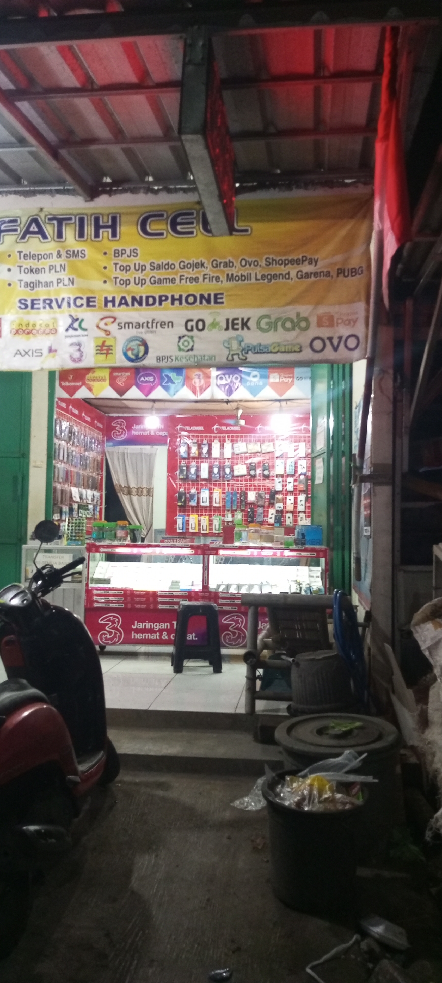 The image shows a brightly lit storefront at night, identified by a banner as "Fatih Cell" and "SERVICE HANDPHONE". The banner lists various services including "Telepon & SMS", "Token PLN", "Tagihan PLN", "BPJS", and top-up services for Gojek, Grab, Ovo, ShopeePay, and games like Free Fire, Mobil Legend, Garena, and PUBG. The storefront displays logos of mobile carriers and payment services such as Indosat Ooredoo, Axis, 3 (Three), Smartfren, Gojek, Grab, Ovo, and Dana.
Inside the store, display cases are filled with mobile phones and accessories. A red scooter is parked to the left of the entrance, and a black stool is placed in front of the display. To the right, there are a few chairs and trash bins, suggesting a slightly cluttered outdoor area. The scene appears to be on a street in Setu, Indonesia, given the context of the store's services and common payment methods in the region. The overall impression is of a local, functional business operating in an urban or semi-urban environment.