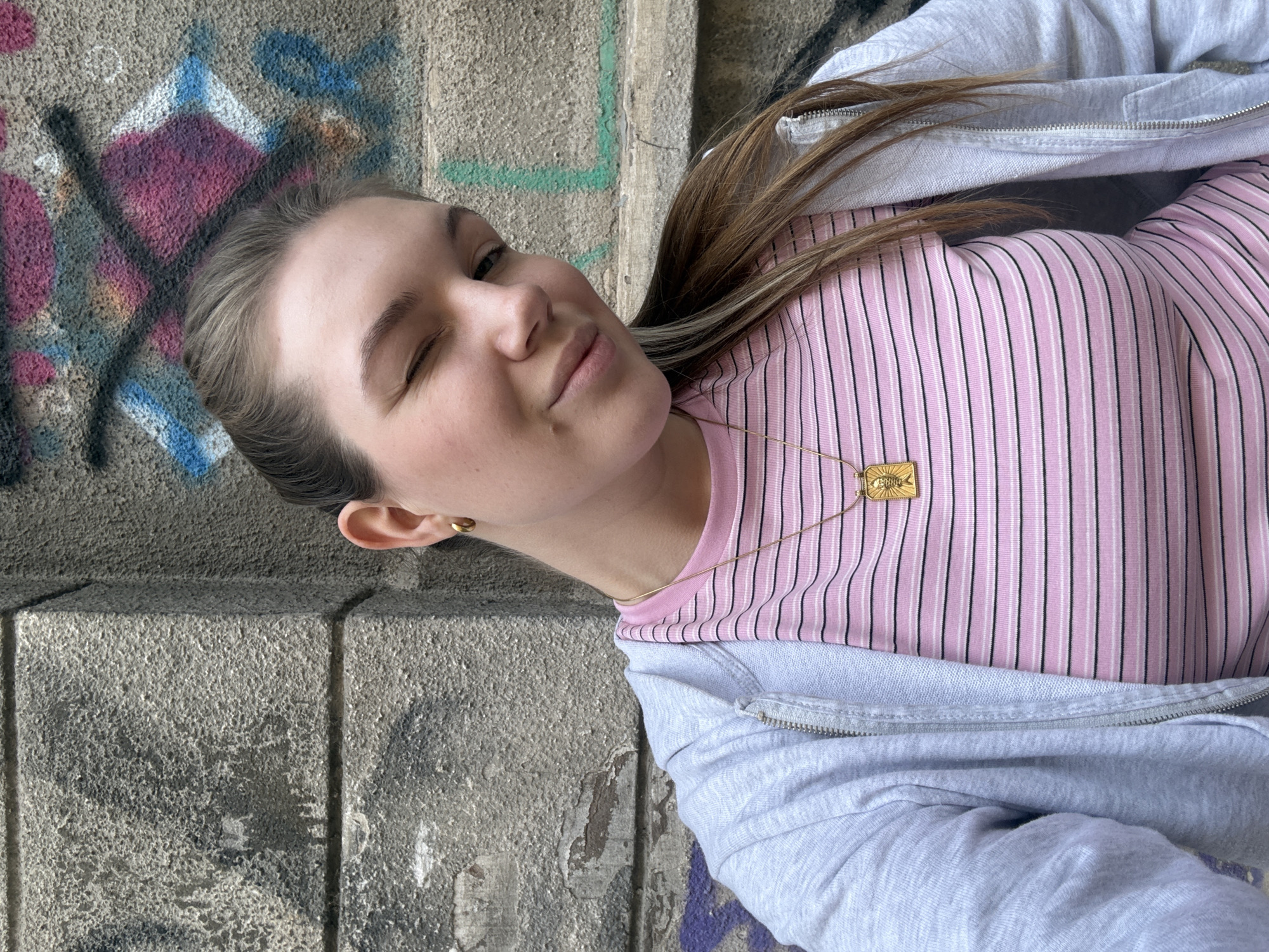 A young woman winks playfully at the camera, posing against a textured concrete wall marked with colorful graffiti. Her head is tilted to the left, and her body extends to the right in the rotated image. She has light brown hair and a fair complexion.
She is dressed in a light pink top with thin black and white vertical stripes, layered under a light grey zip-up hoodie. A delicate gold chain necklace with a rectangular pendant featuring a stylized sun design and the faint engraved text "PERCHE" is visible around her neck. She wears a small gold stud earring in her visible ear.
The concrete background shows signs of wear and various patches of abstract graffiti in shades of pink, blue, white, and black, including cross and diamond-like shapes. The lighting suggests it is daytime. The woman's expression is casual and cheerful. The setting is an urban concrete area, consistent with locations found in Belgrade, Serbia.